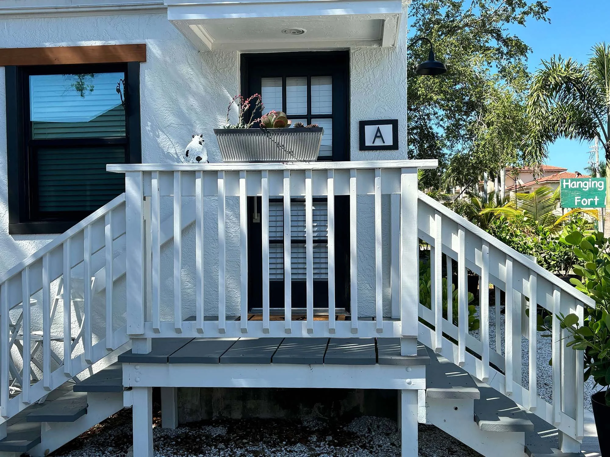 White house with black-framed windows and a staircase with white railing, decorated with a cow figurine and a planter on the porch, surrounded by greenery and trees, with a sign reading 'Hanging Fort' in the background.