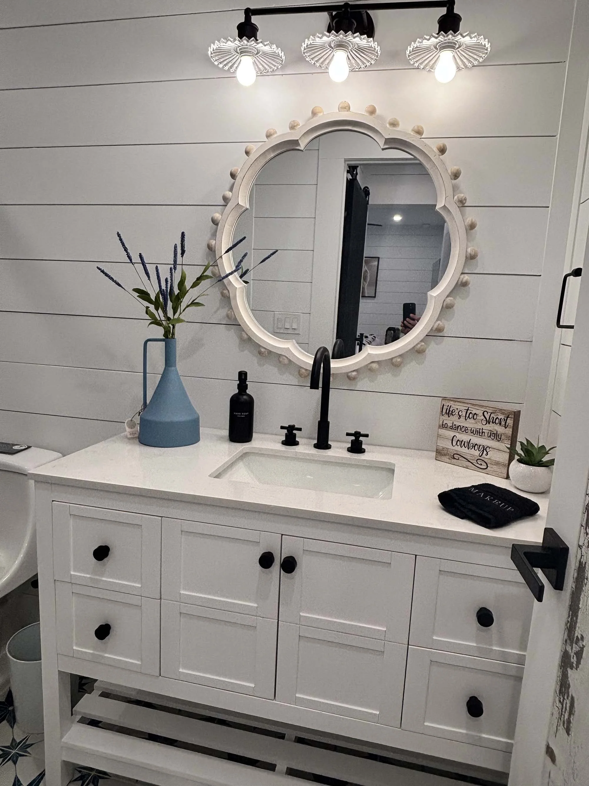 Modern white bathroom vanity with black knobs, a black faucet, a large decorative mirror with a white frame and round accents, a blue vase with tall lavender plants, a black soap dispenser, a small wooden sign with quotes, a white potted succulent, a