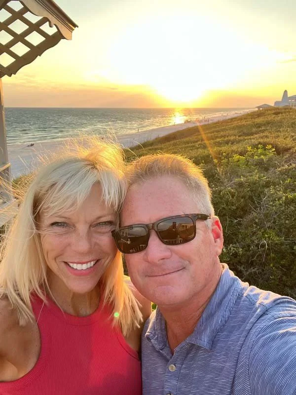 A smiling woman and man taking a selfie at the beach during sunset, with the ocean, green beach vegetation, and a pier in the background.