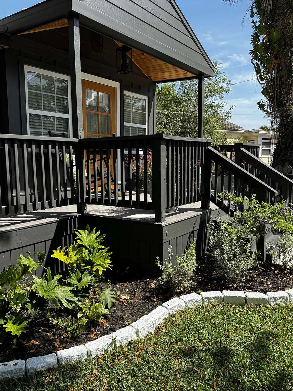 Back porch of a house with black railing, stairs, and a wooden deck. Green plants and shrubs in a landscaped garden bed in front.