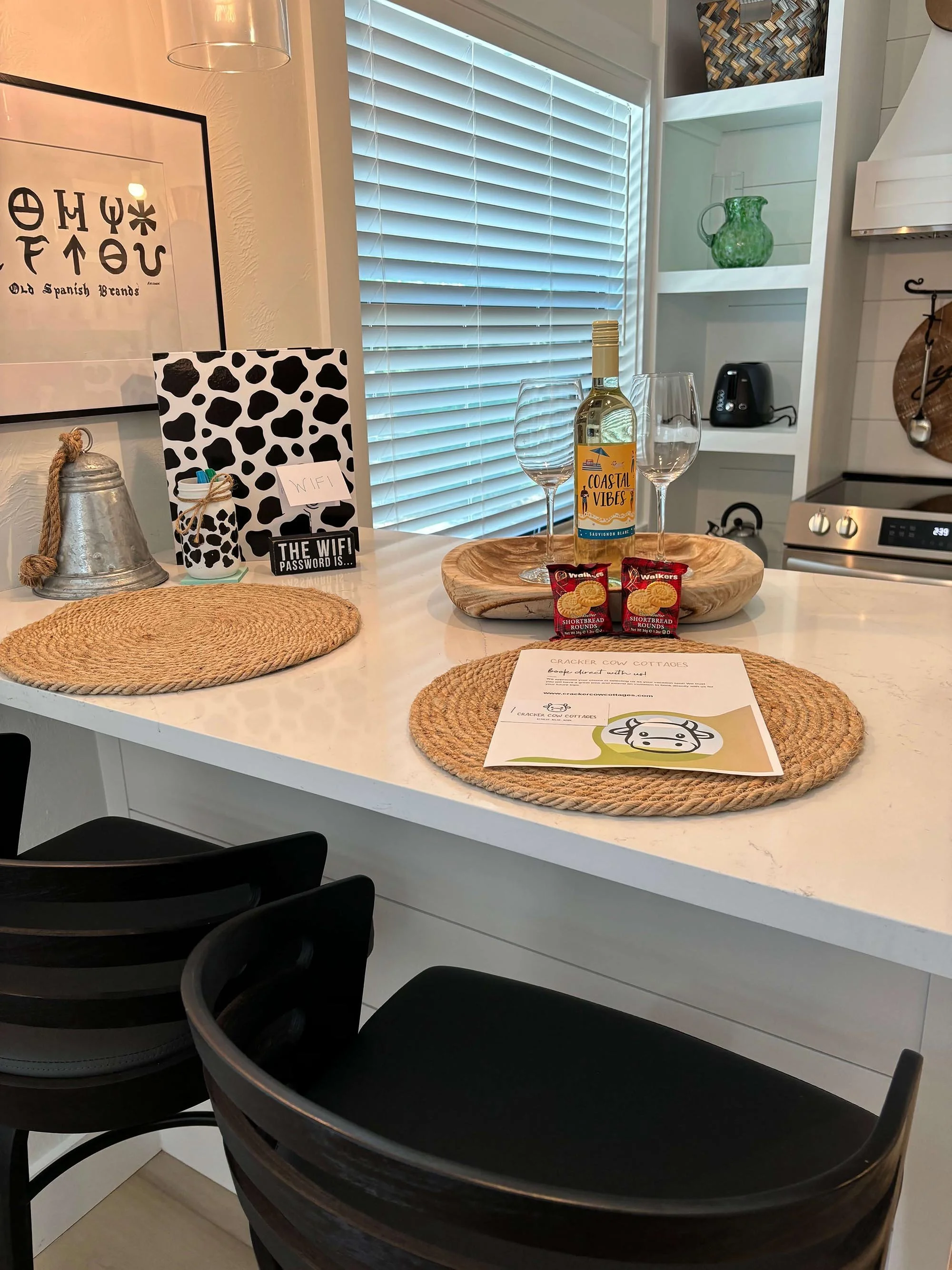 Kitchen countertop set for wine and snacks, with two wine glasses, a bottle of wine labeled 'Coastal Vibes,' two packets of crackers, and a wooden bowl. There are decorative placemats, a framed sign with symbols, and a small tray with a chalkboard la