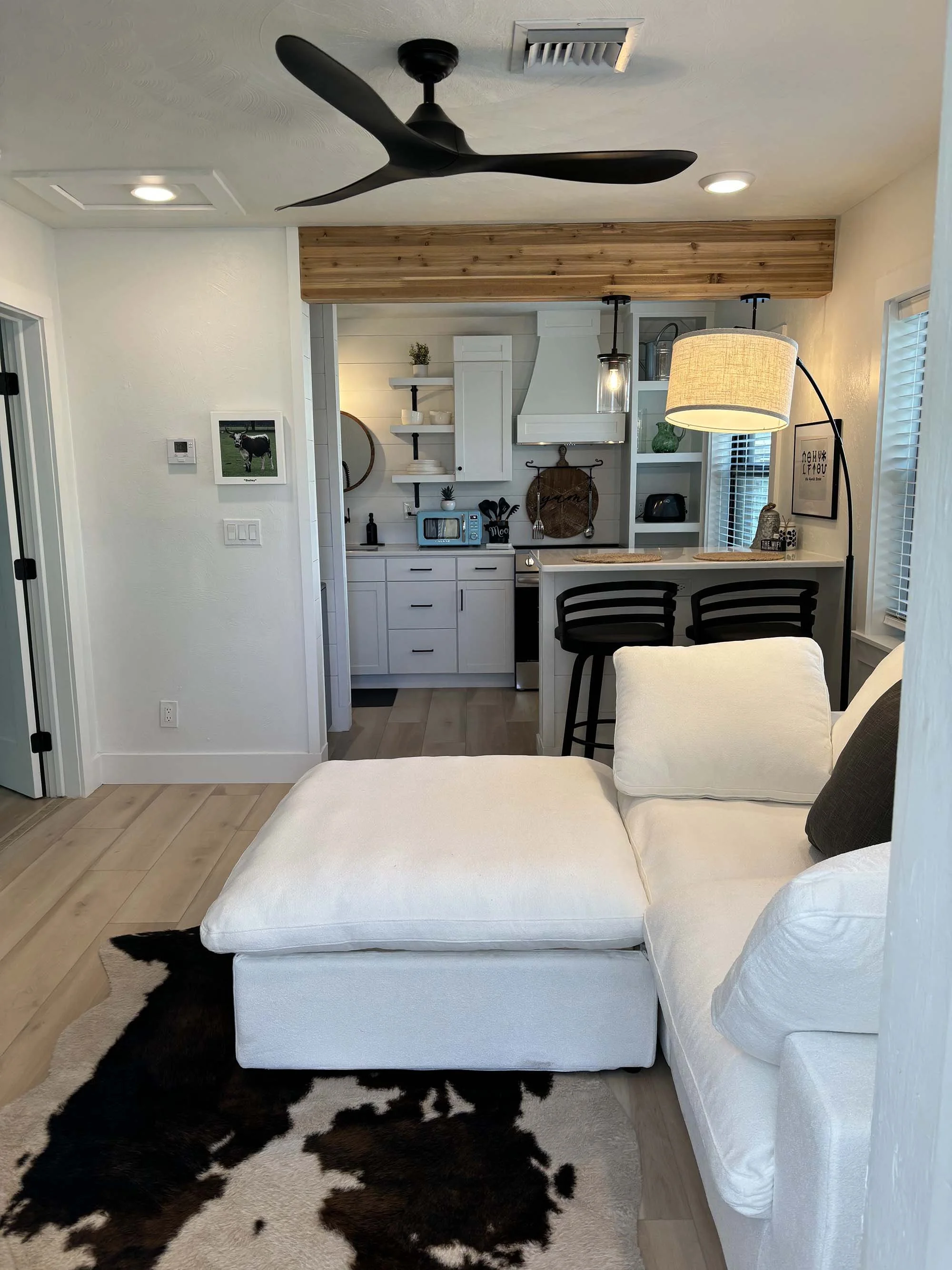 Open-concept living room with white sectional sofa, cowhide rug, and kitchen in the background with white cabinets, black accents, and a small breakfast bar with two black chairs.