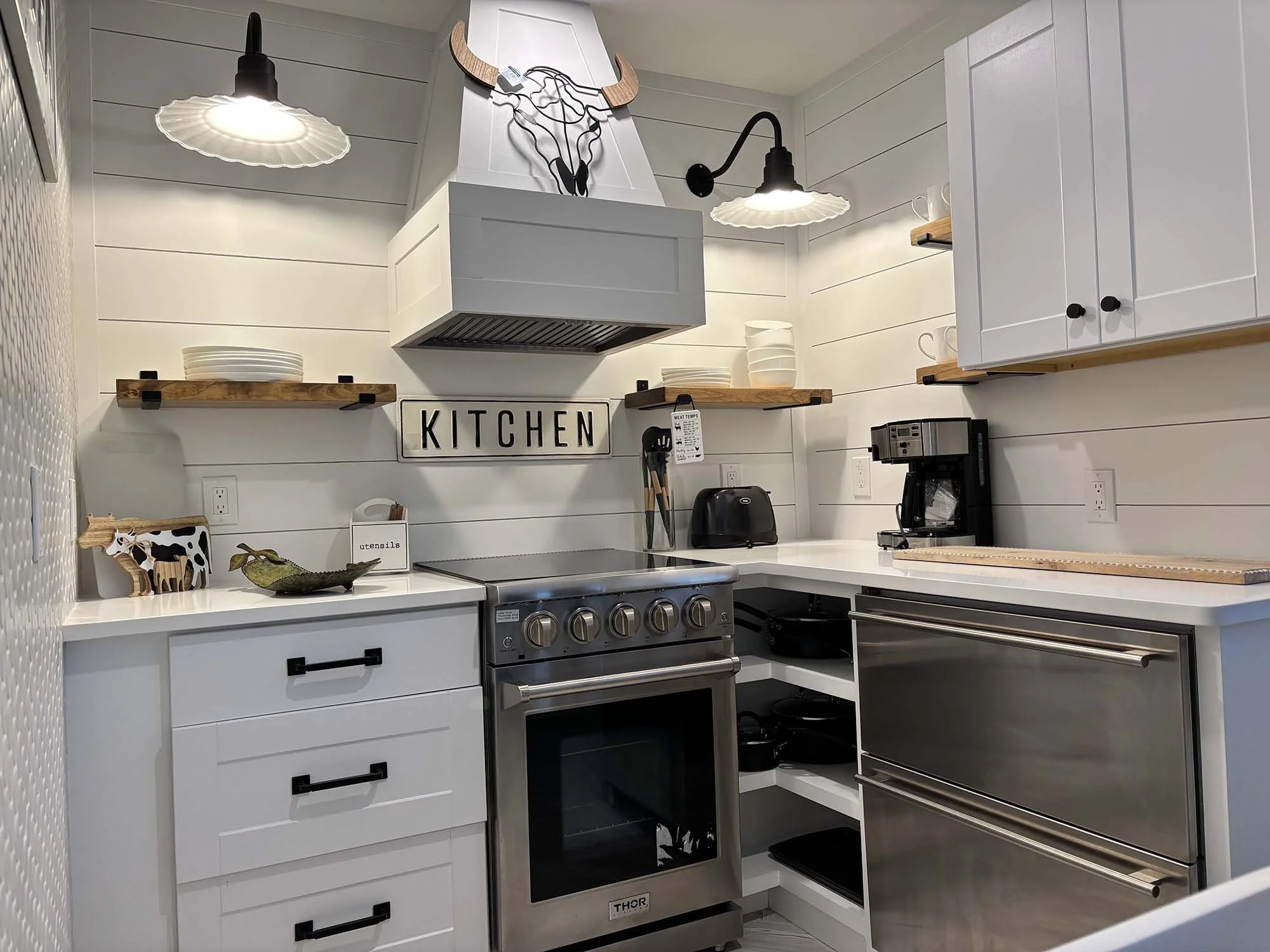 A small kitchen with white cabinets, a stainless steel oven, a coffee maker, and wooden shelves holding dishes and decorative items. There is a black light fixture and a wall art piece of a cow's head above the range.