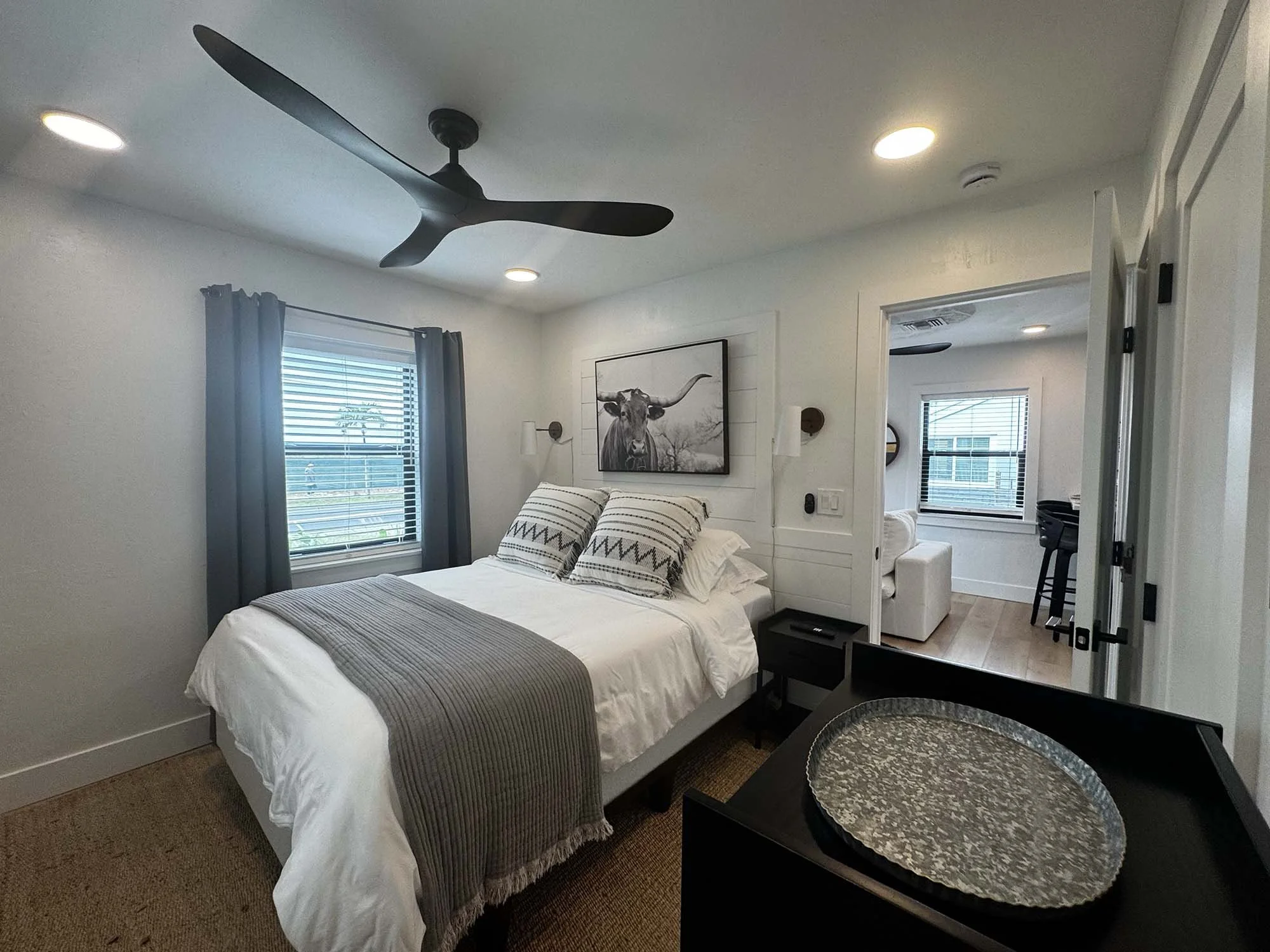 A bedroom with a white bed, striped pillows, and a gray throw blanket. There's a window with gray curtains, a framed black-and-white bull photograph on the wall, and a ceiling fan. The room has a nightstand, a small sitting area with a white armchair