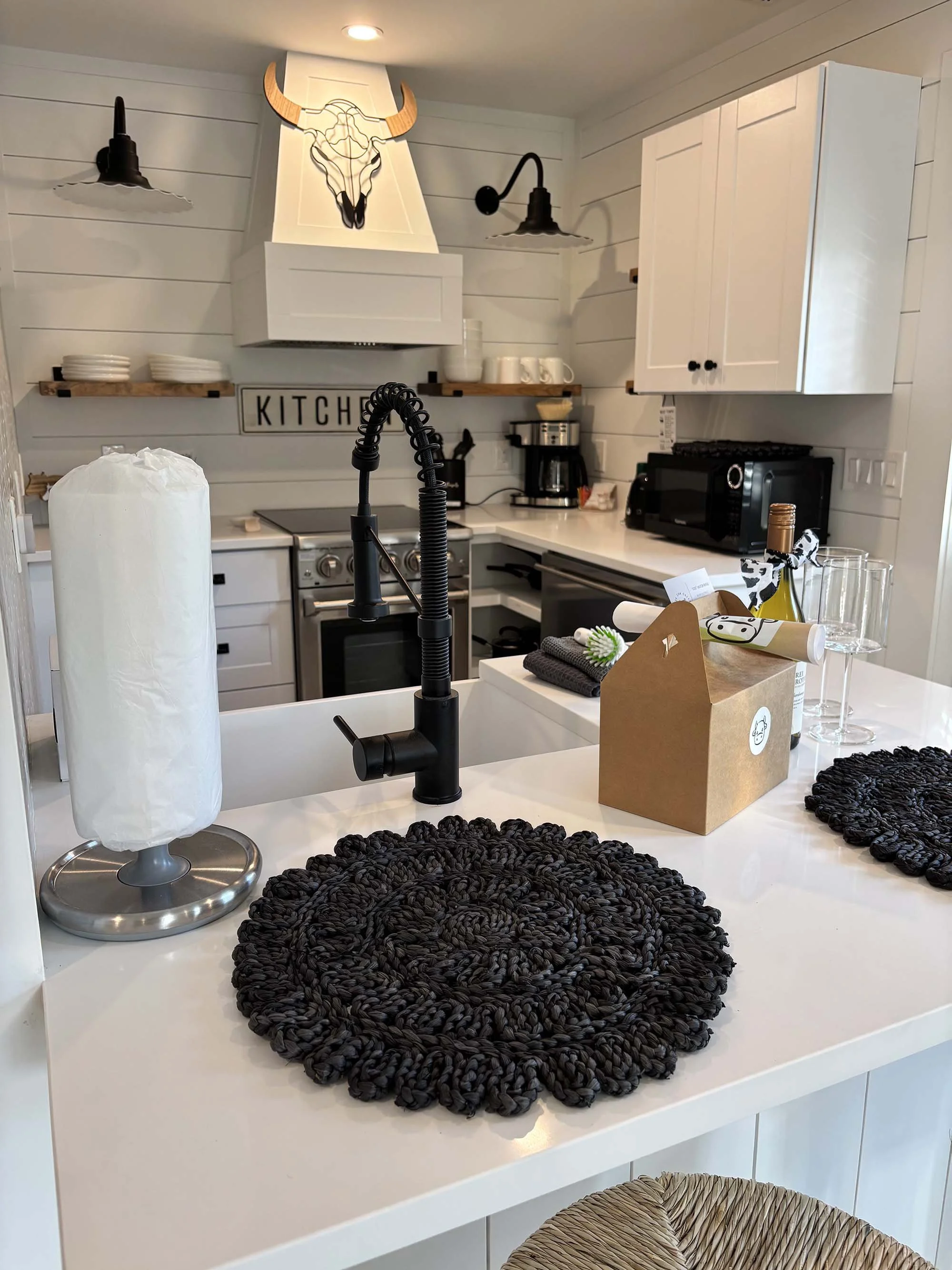 Modern kitchen with white shiplap walls, black accents, a black matte faucet, and a countertop with a round black woven placemat. The kitchen features a white range hood with a decorative animal skull and horns art piece above it, and black sconces. 
