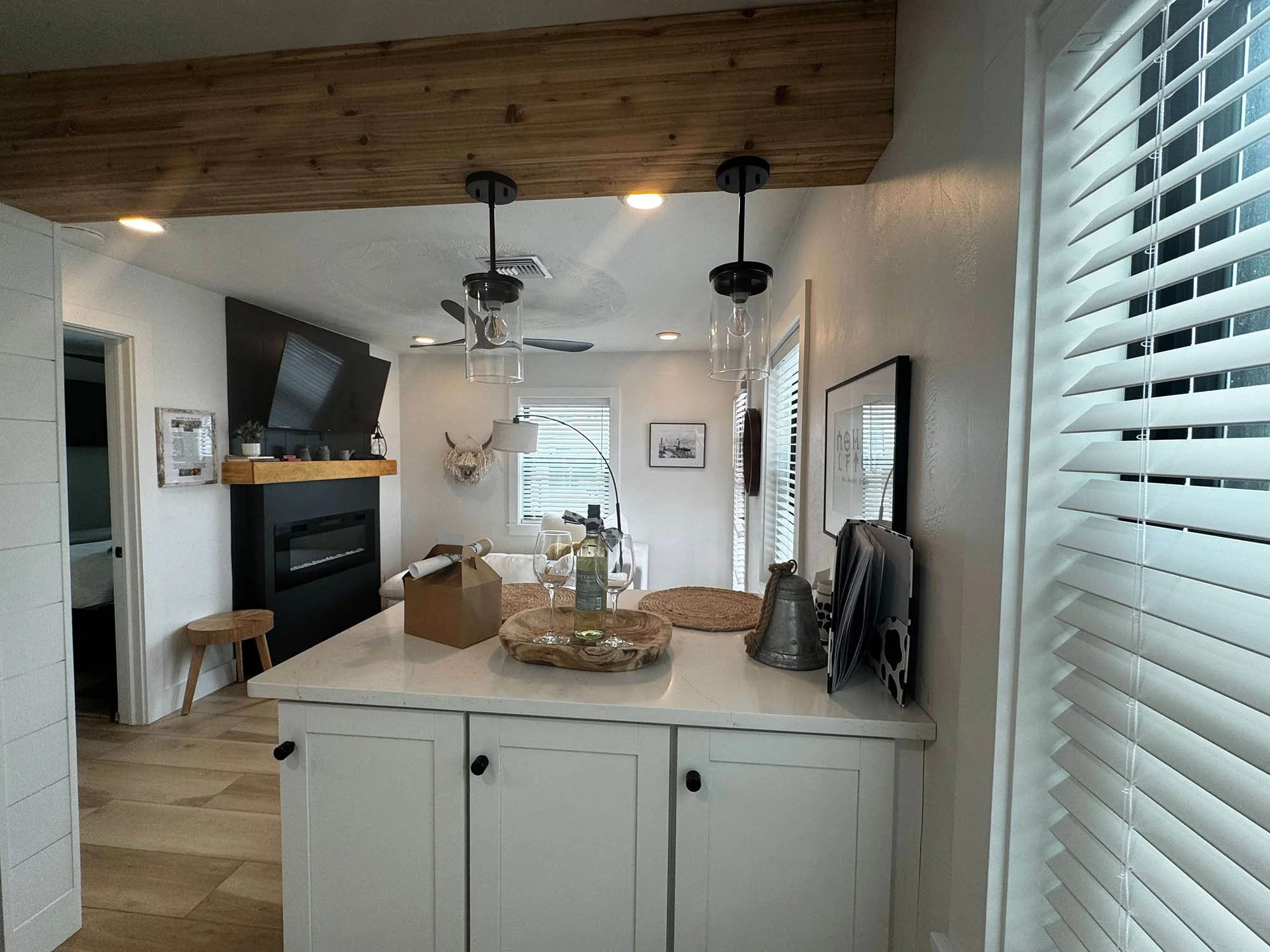 Modern living room with a white kitchen island, pendant lights, large windows with blinds, a white sofa, a black fireplace, and wall-mounted TV, decorated with wall art, a basket, and a floor lamp.