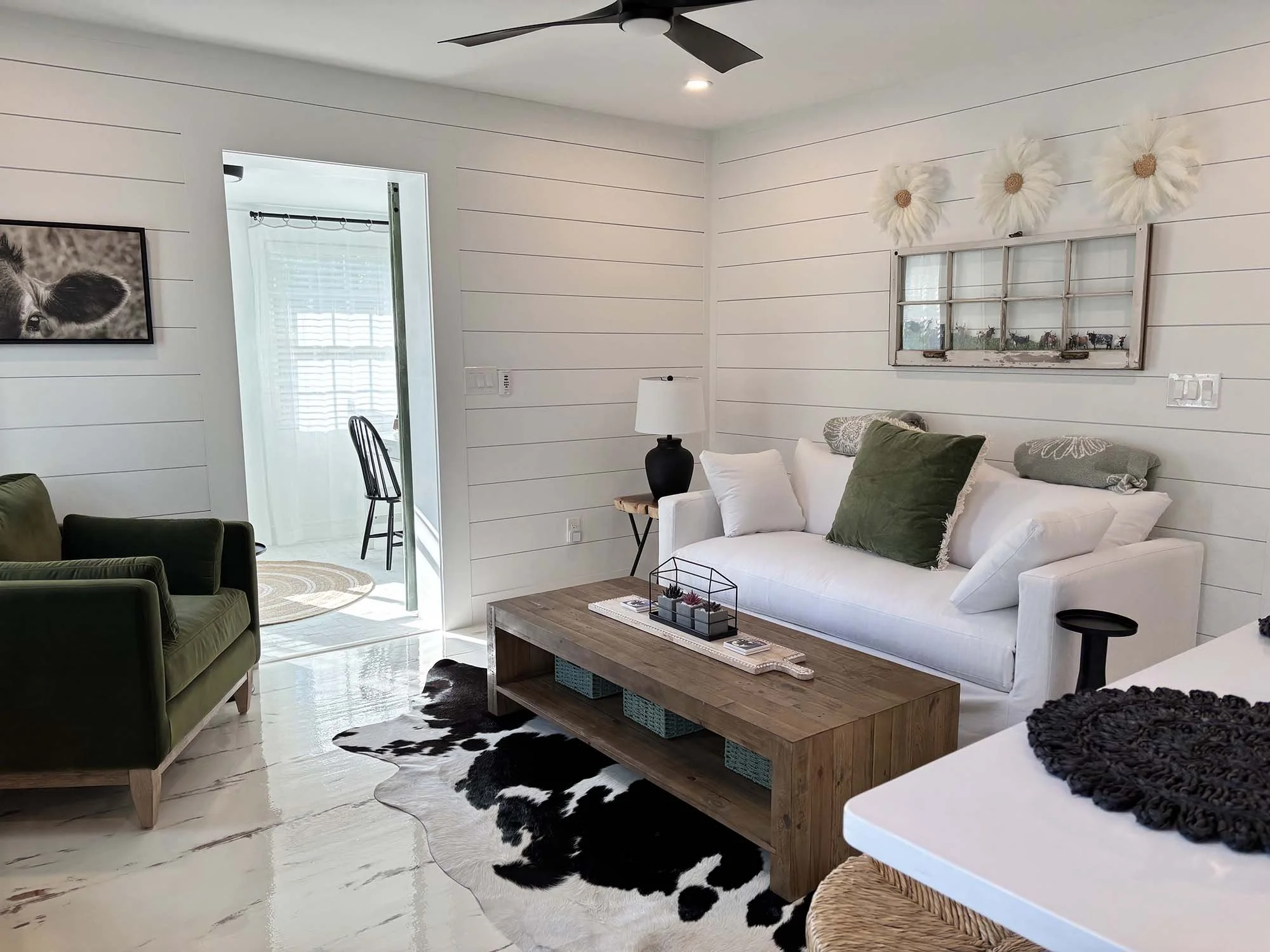 Living room with white shiplap walls, a white sofa with throw pillows, a wooden coffee table, a cowhide rug, dark green armchair, and wall décor including a framed animal photo and white feathered flowers.