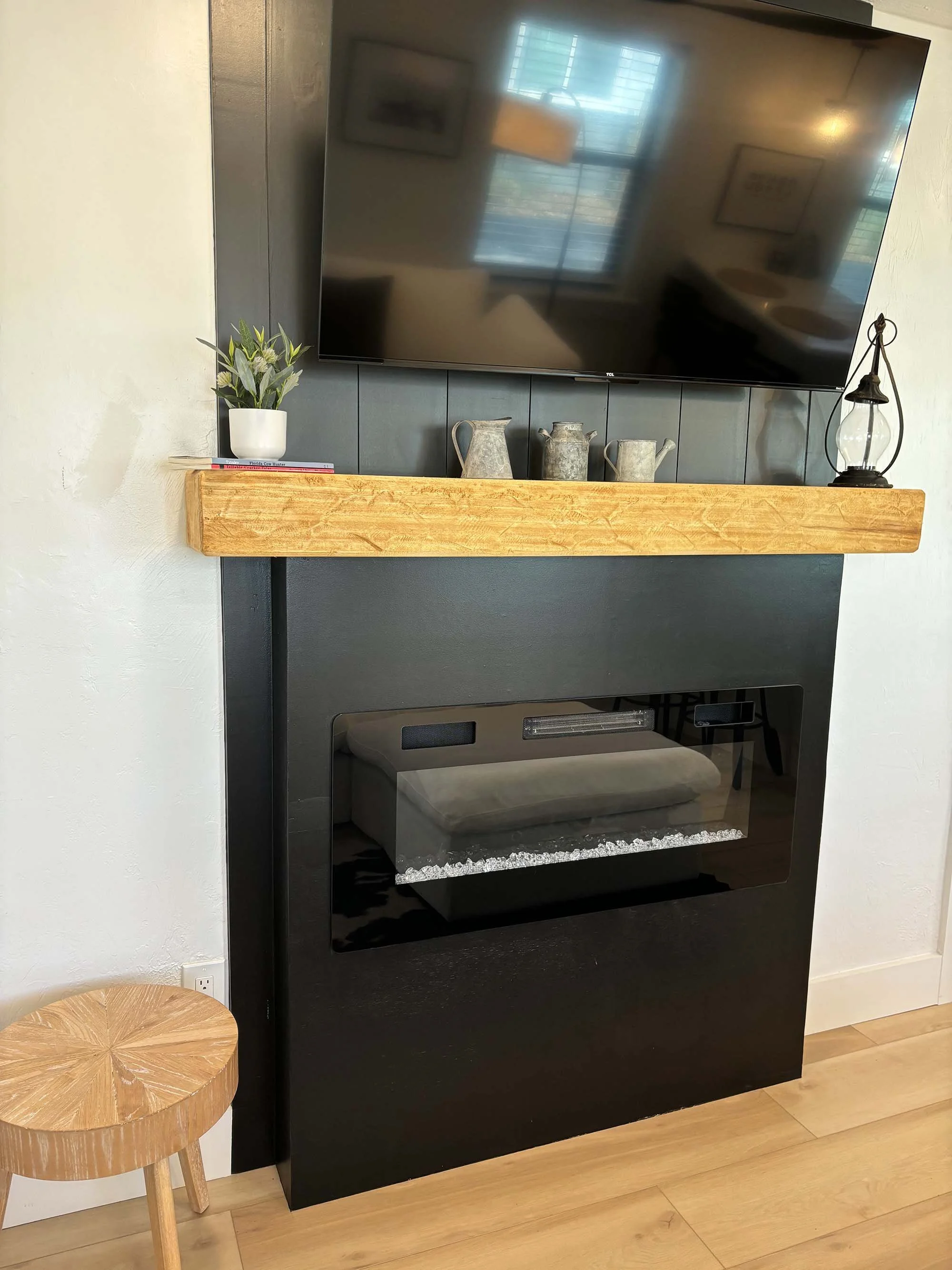 Living room fireplace with a mounted flat-screen TV above, decorated with ceramic pitchers, a small potted plant, and a lantern on the mantle. There is a small wooden stool beside it.