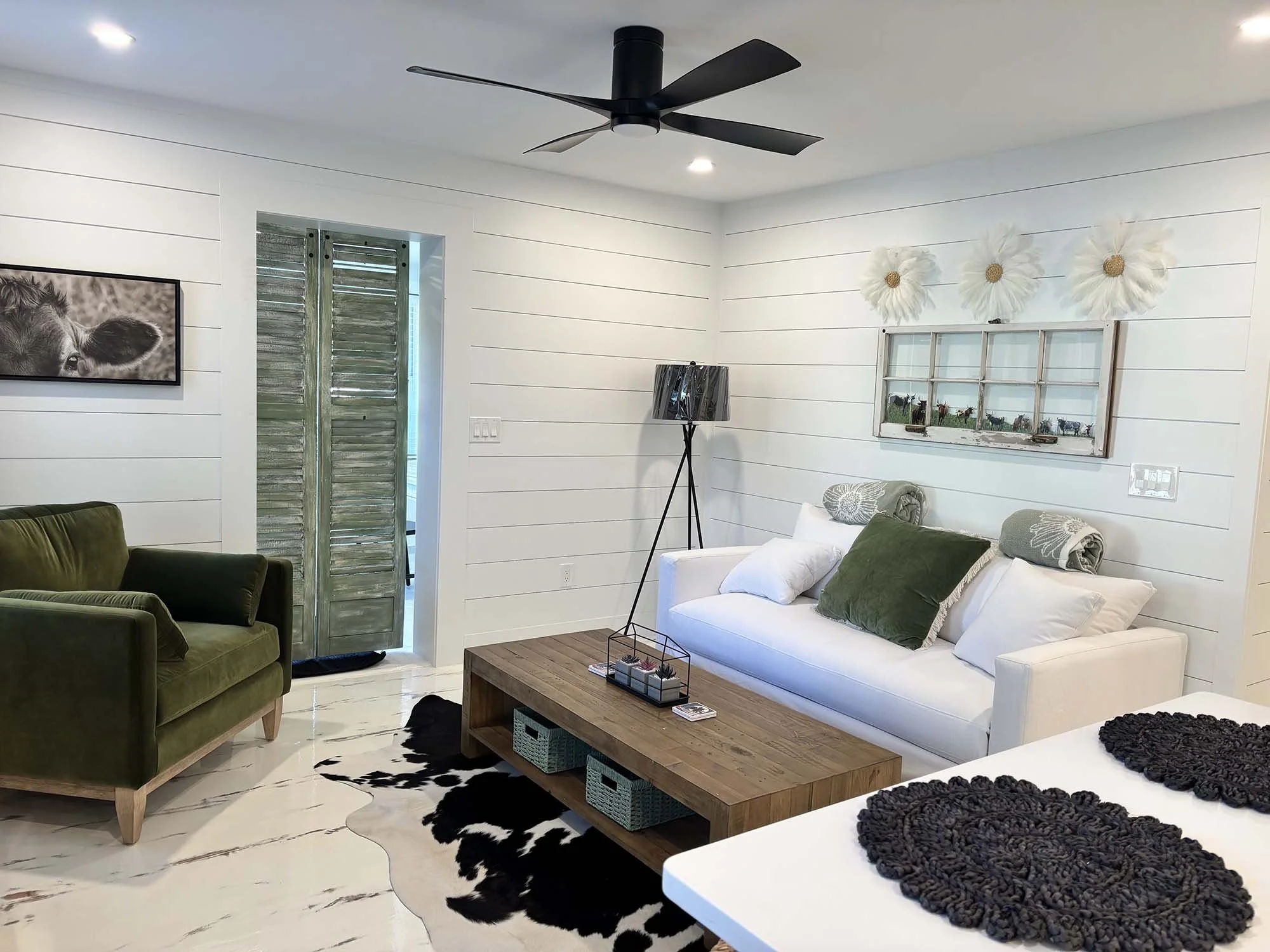 Living room with white shiplap walls, a white sofa with green and decorative pillows, a wooden coffee table, an green armchair, cowhide rug, black floor lamp, framed wall art, ceiling fan, and window with wooden shutters.