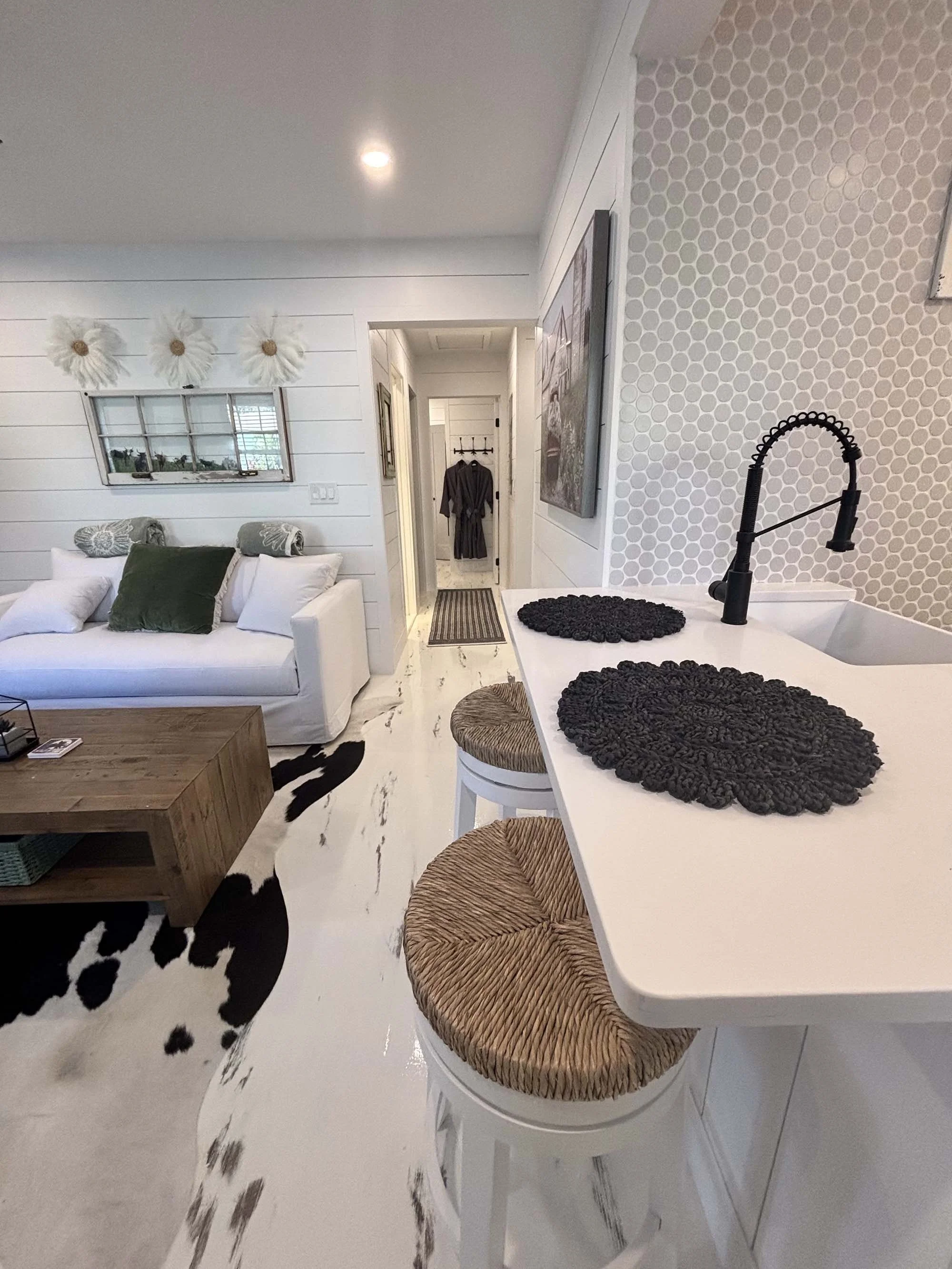 Living room with white sofa, wooden coffee table, wall decor, and a kitchen counter with black textured placemats and a black faucet, decorated in neutral and black tones.
