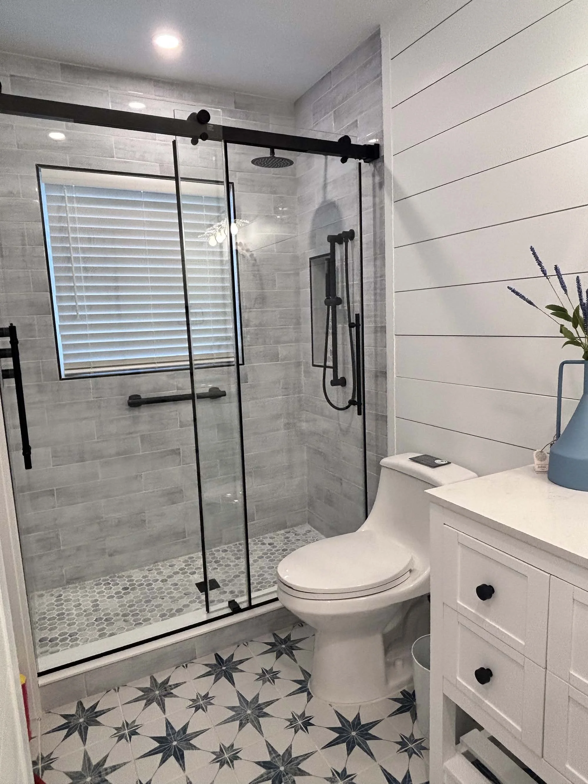 Modern bathroom with a walk-in shower with glass sliding doors, gray tiled walls, and a window with white blinds, a white toilet, and a white cabinet with black knobs topped with a blue vase and purple flowers.