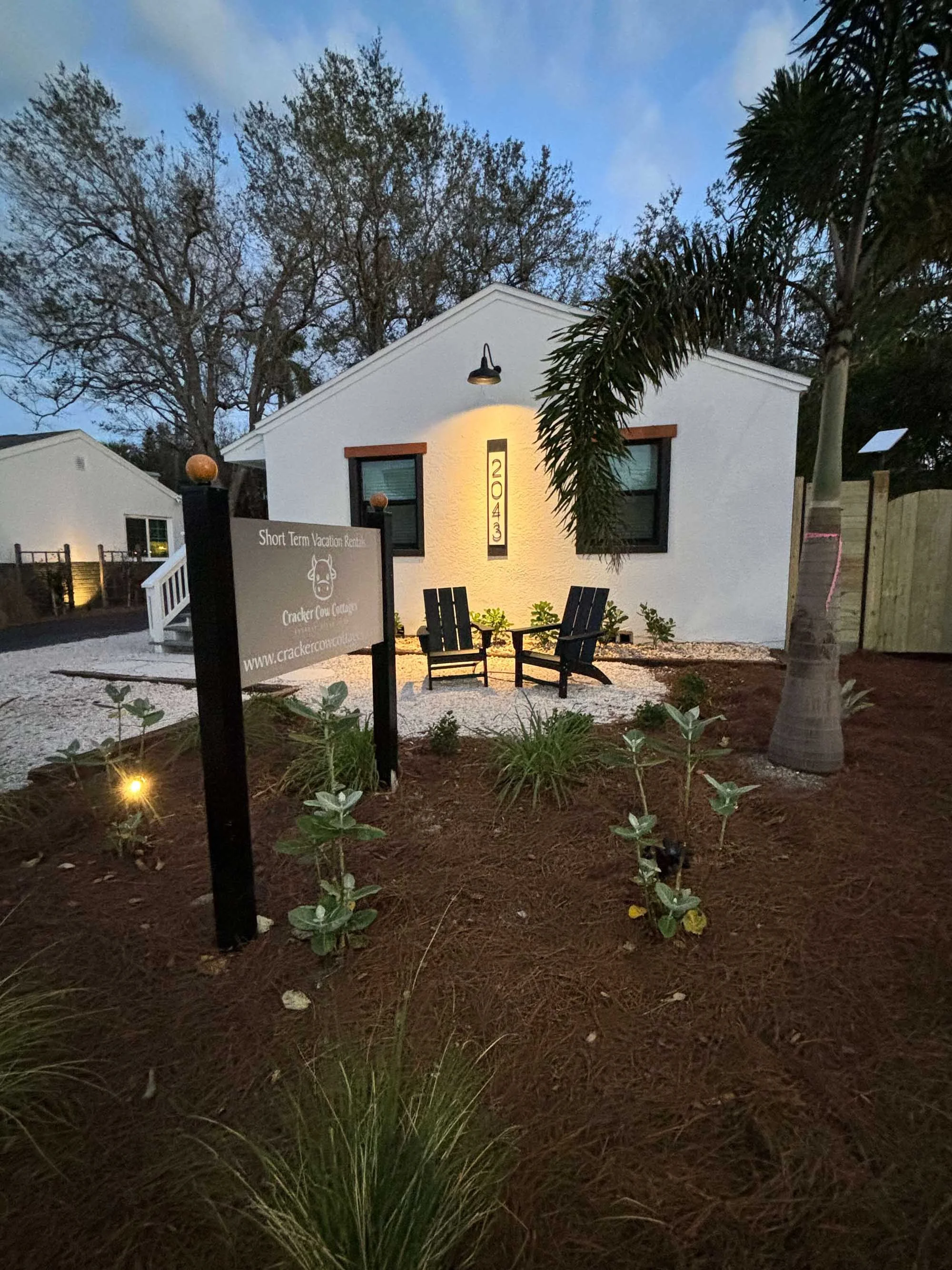 Small white building with black-framed windows, surrounded by landscaped garden with young plants and a palm tree. There are two black chairs and a sign that reads "Short Term Vacation Rentals - Cracker Cow Cottage." The building's address is 2043.
