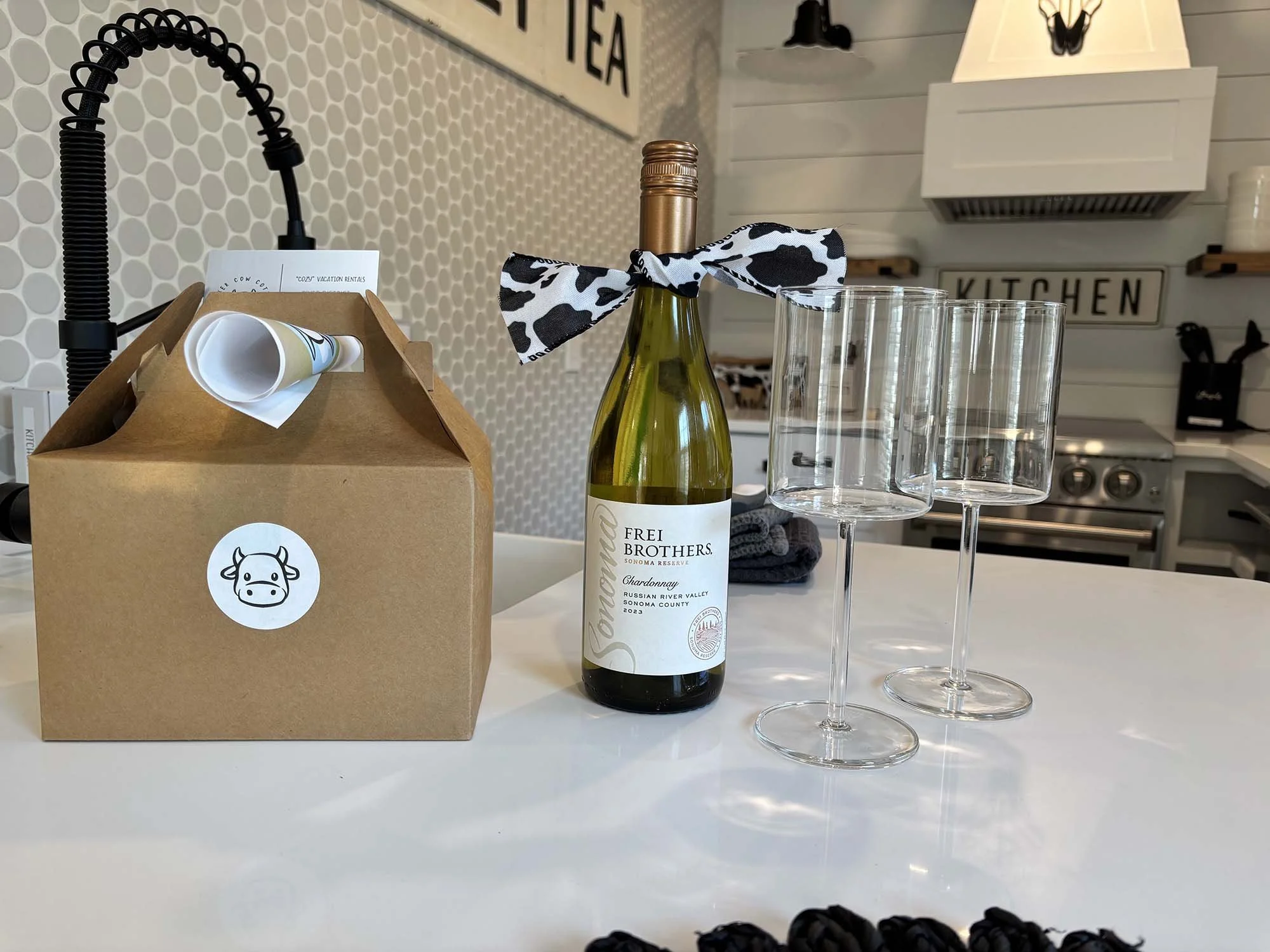 A white kitchen counter with a bottle of wine with a black and white cow print ribbon, three empty wine glasses, a brown pizza box with a cow logo, and a kitchen background with a stove and shelves.