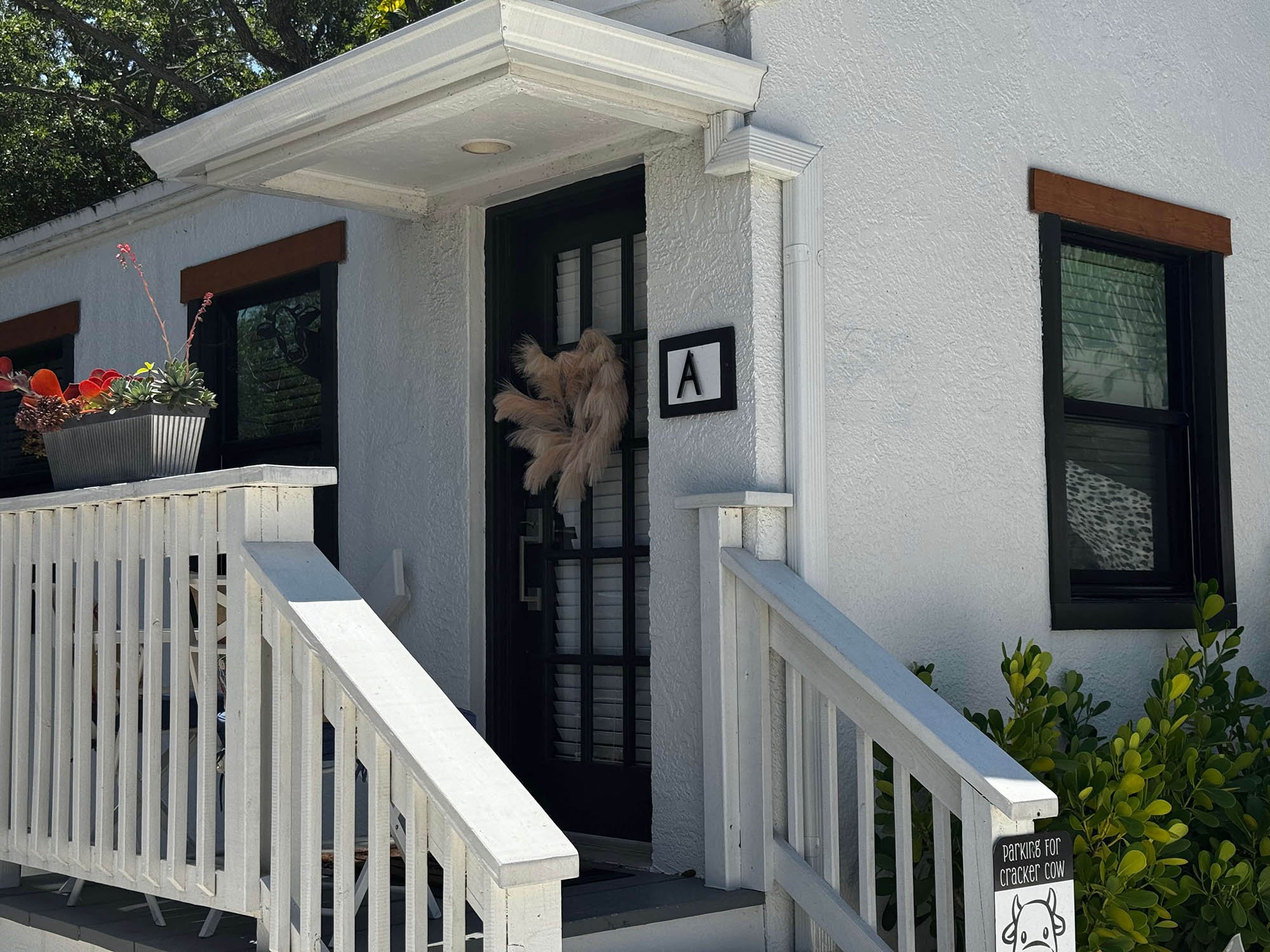 White house with black door, a decorative wreath on the door, window boxes with plants, and a sign that reads "Parking for cracker cow" near the stairs.