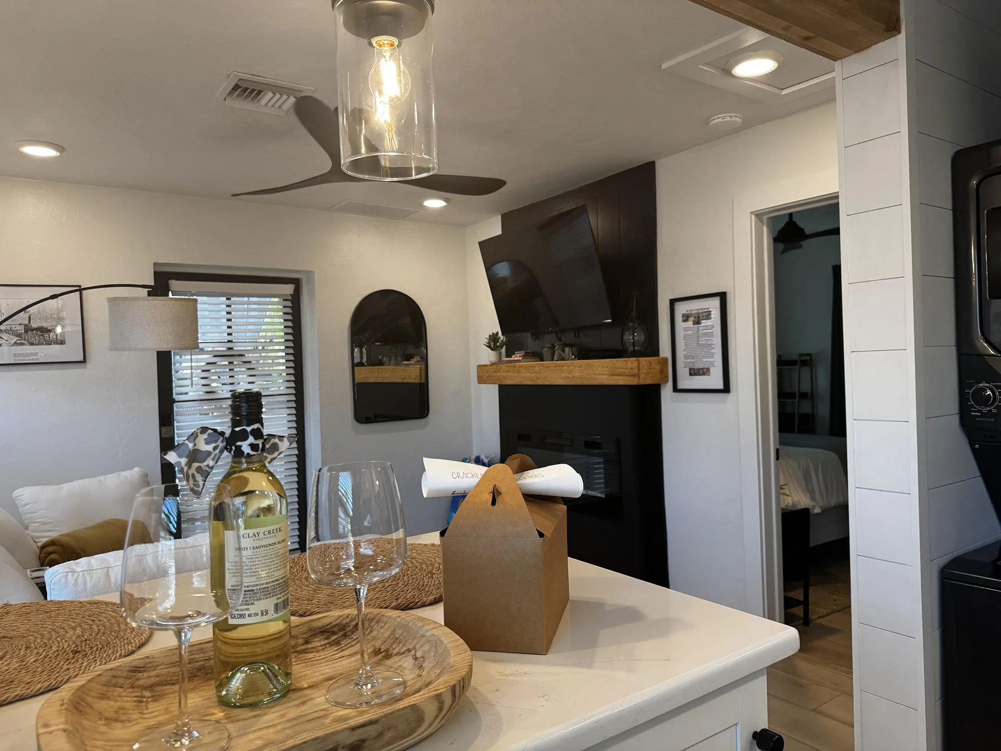 Kitchen and living room area with a white counter, wine bottle, two empty wine glasses, and a small brown paper bag on the counter. In the background, a wall-mounted TV above a fireplace, a mirror, a window with blinds, a lamp, and a ceiling fan are 
