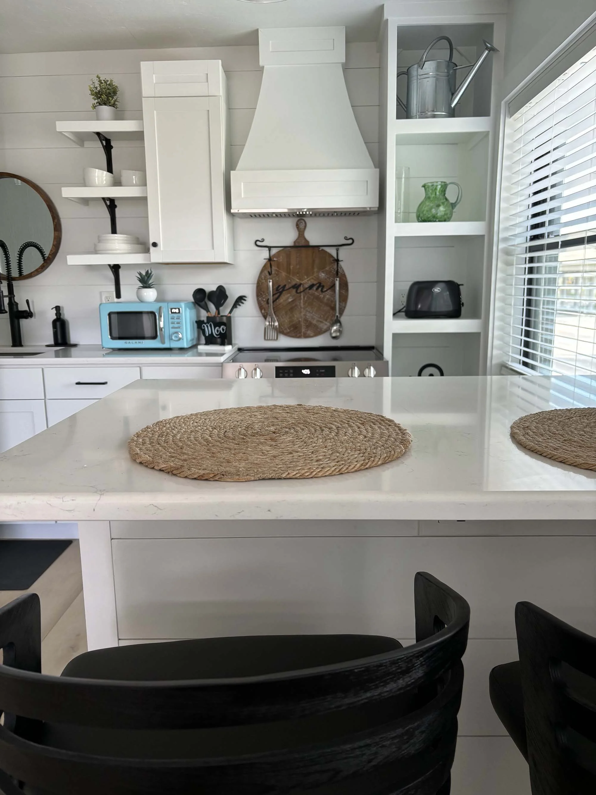 Modern white kitchen with a marble island, black chairs, woven placemats on the countertop, white cabinets, and open shelving with decorative items, a microwave, and a toaster, along with a window with blinds.