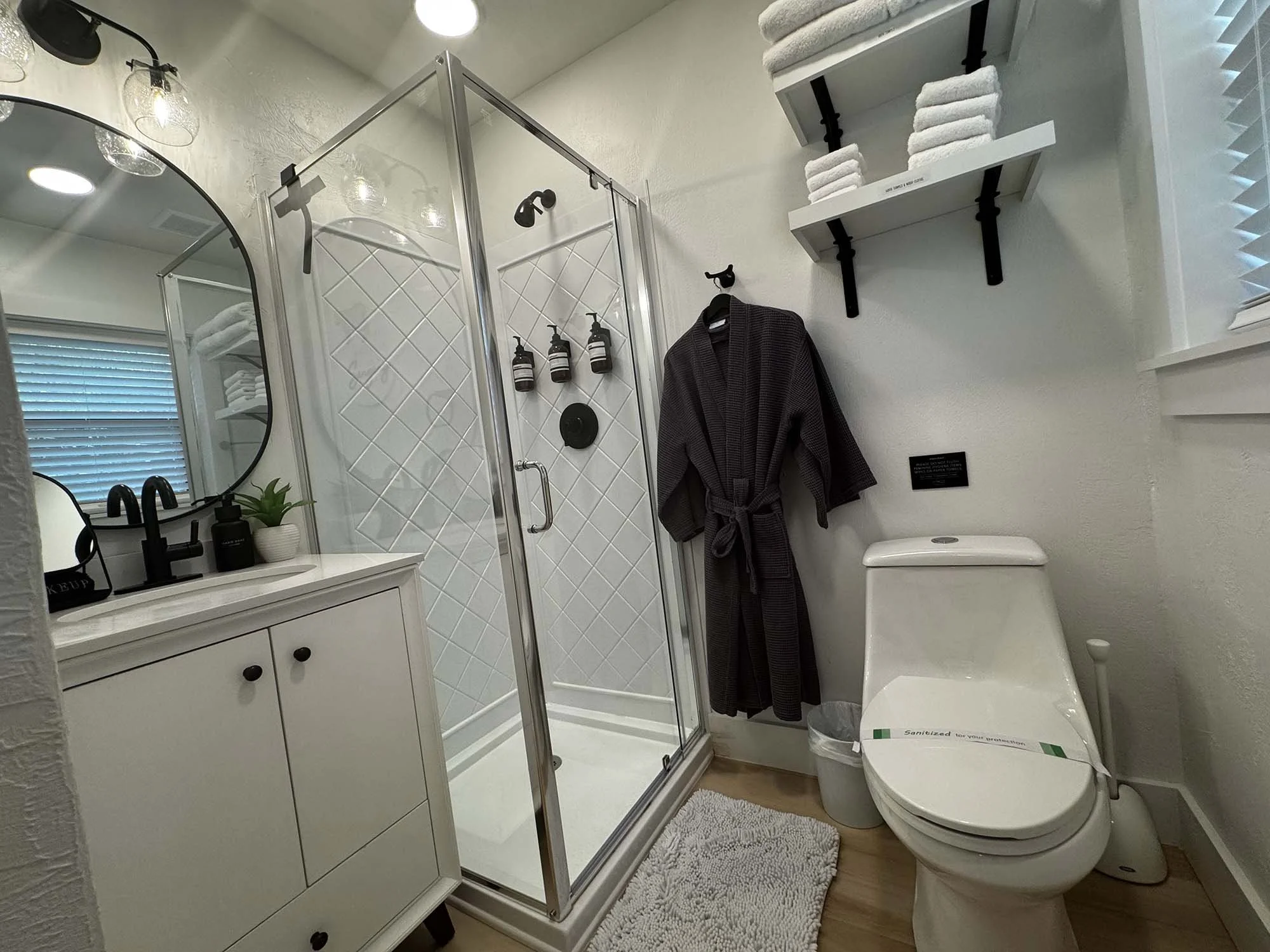 A modern bathroom with a glass shower enclosure, white vanity with a round mirror, black fixtures, a black robe hanging on a wall hook, and a toilet with shelves above holding folded white towels.