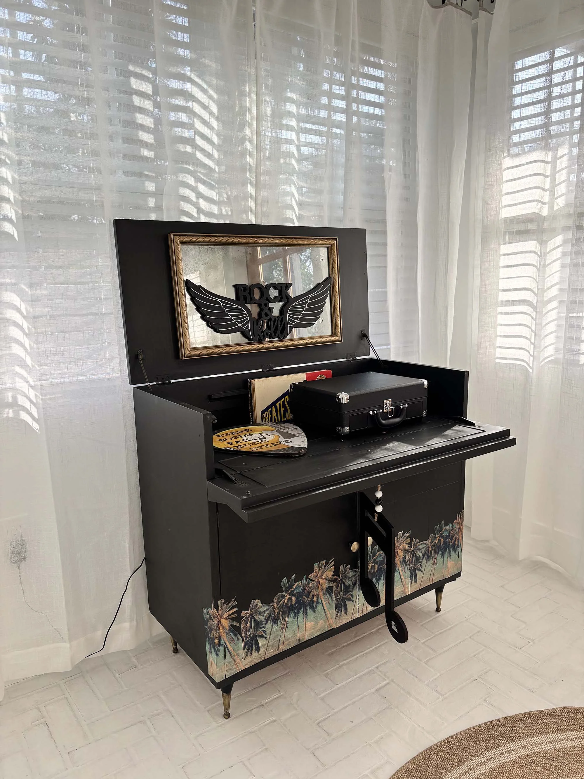 A vintage black piano with a palm tree design on the bottom front, topped with a small case and a record. A mirror on the lid has a 'Rock & Roll' sign with wings attached.