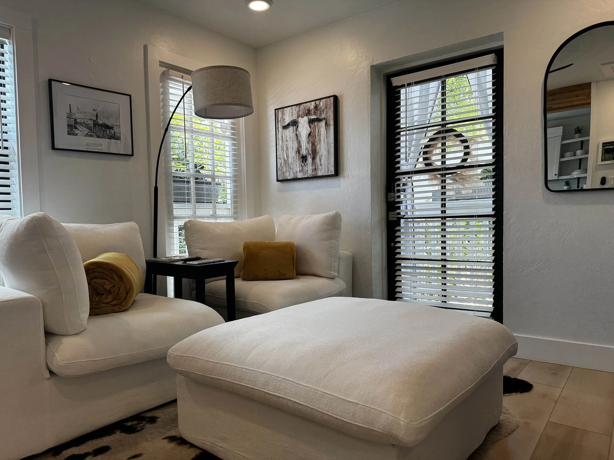 A cozy living room with white couches, a black side table, yellow pillows, a tall floor lamp, framed artwork, windows with blinds, and a door with blinds leading outside.
