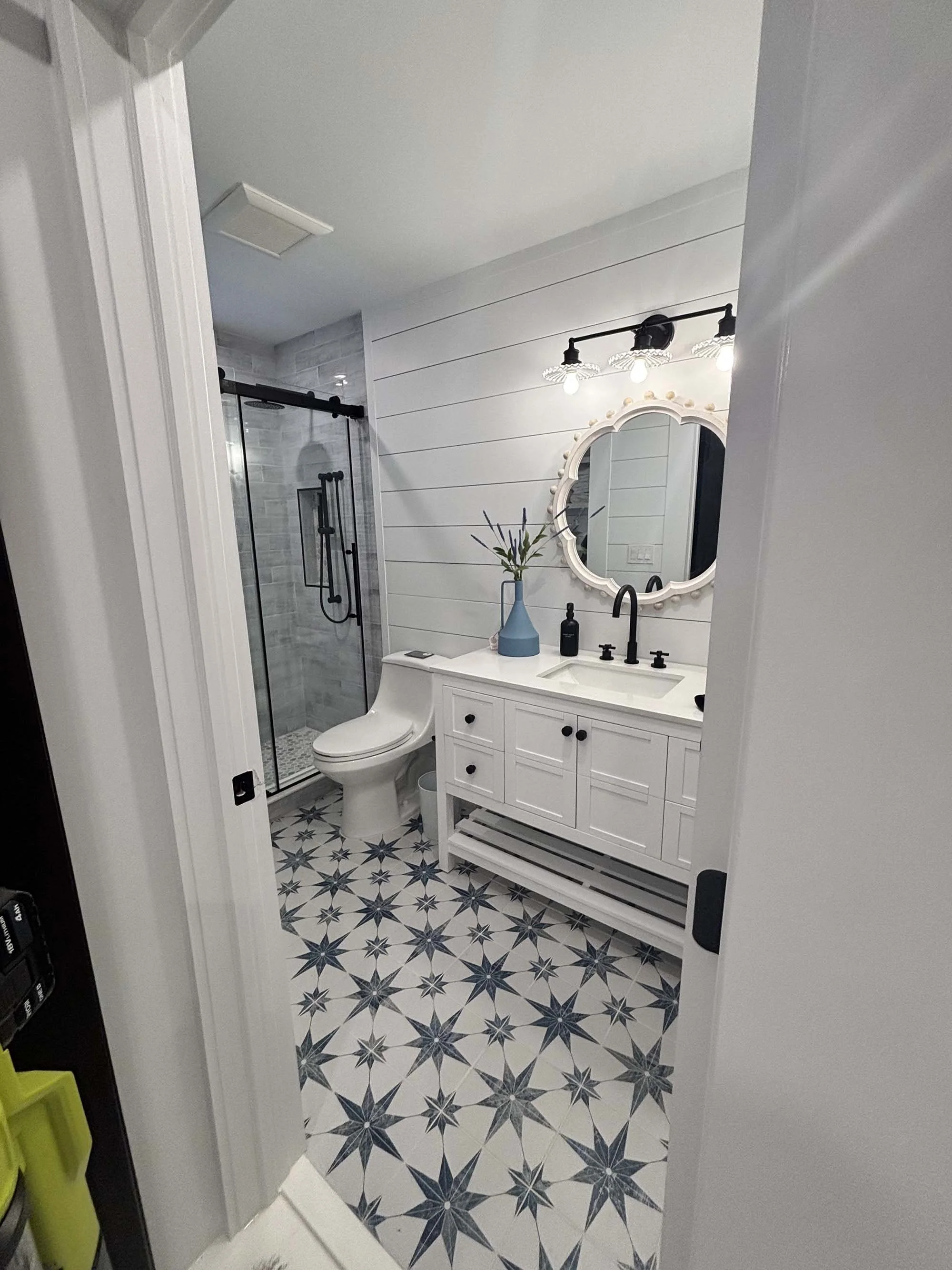 Modern bathroom with a glass shower, white vanity with black fixtures, mirror, and black lighting fixture. Blue vase with green branches on the vanity, patterned star tile floor, and white shiplap walls.