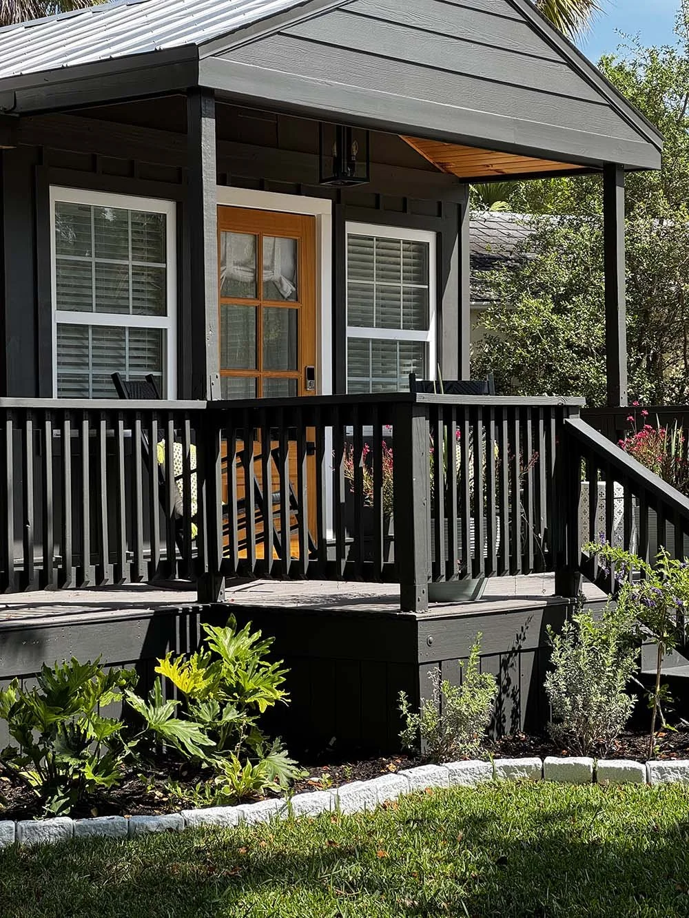 A front porch of a house with black railings, a wooden door with glass panels, and a covered roof. There are chairs on the porch, and the front yard has green plants and a flower bed.