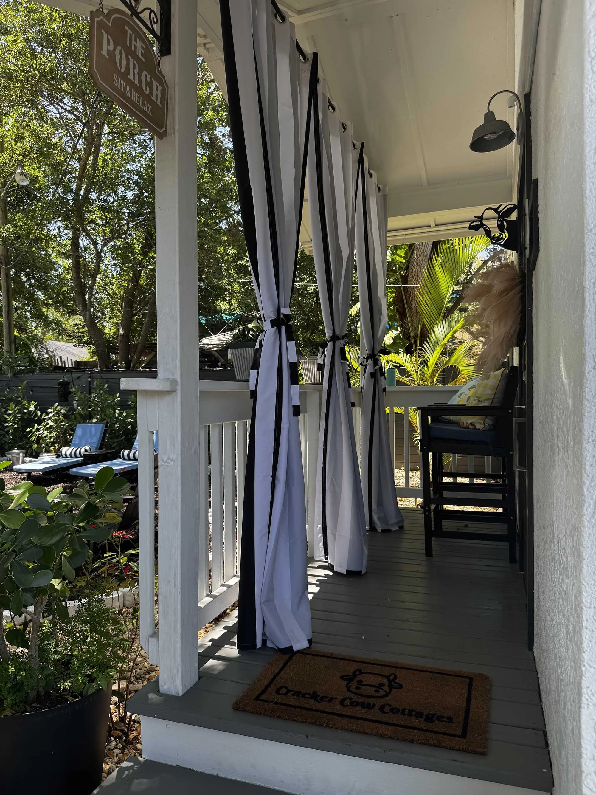 Front porch with white curtains tied to sides, a black chair with a yellow pillow, pampas grass decoration, potted plants, and a welcome mat with a cow face and the text "Cracker Cow Concess".
