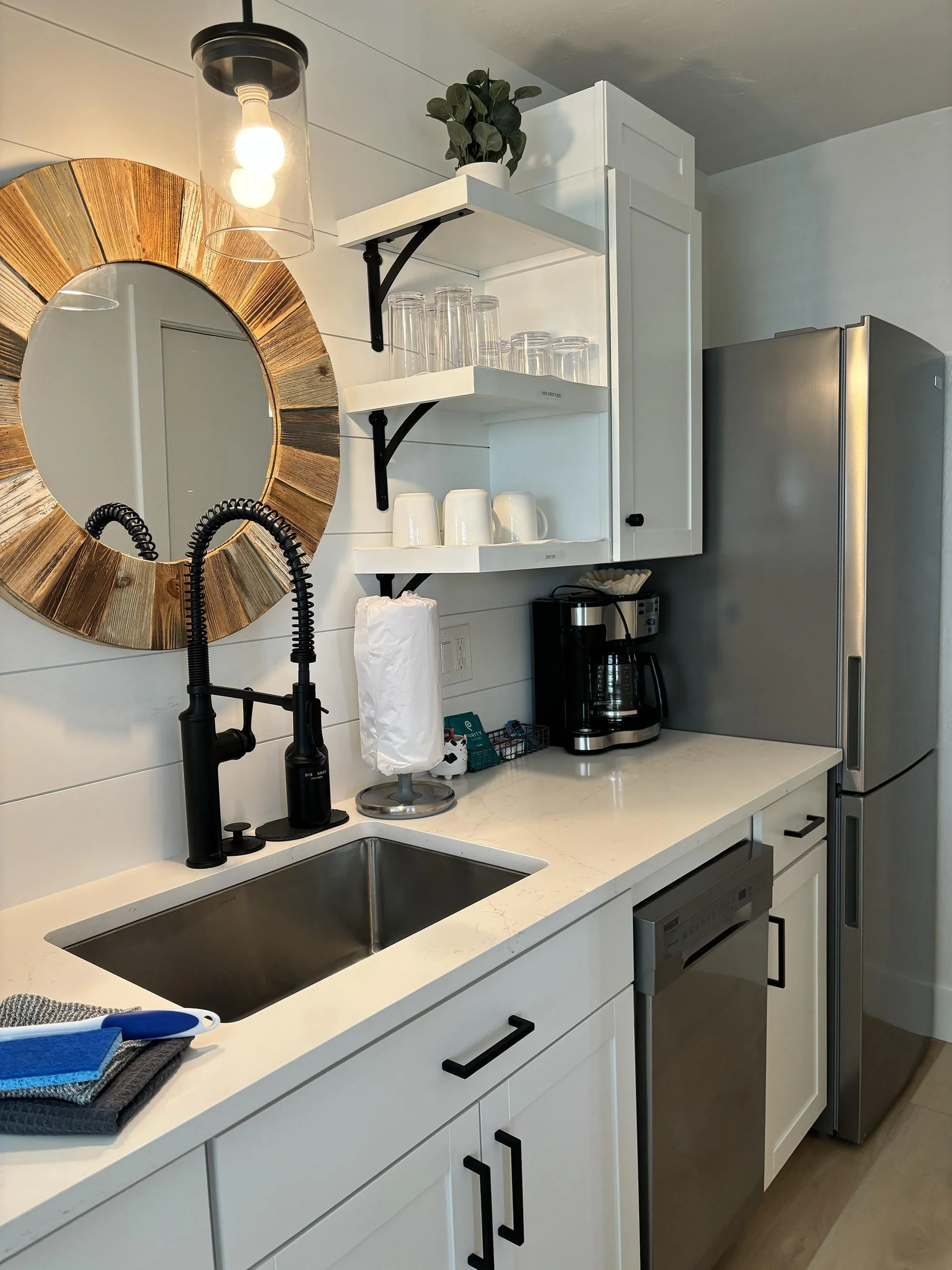 Kitchen with white cabinets, a black faucet over a stainless steel sink, open shelves with glasses and cups, a coffee maker, and a large refrigerator. A round, gold-colored mirror hangs on the wall, and a decorative light fixture with exposed bulbs i