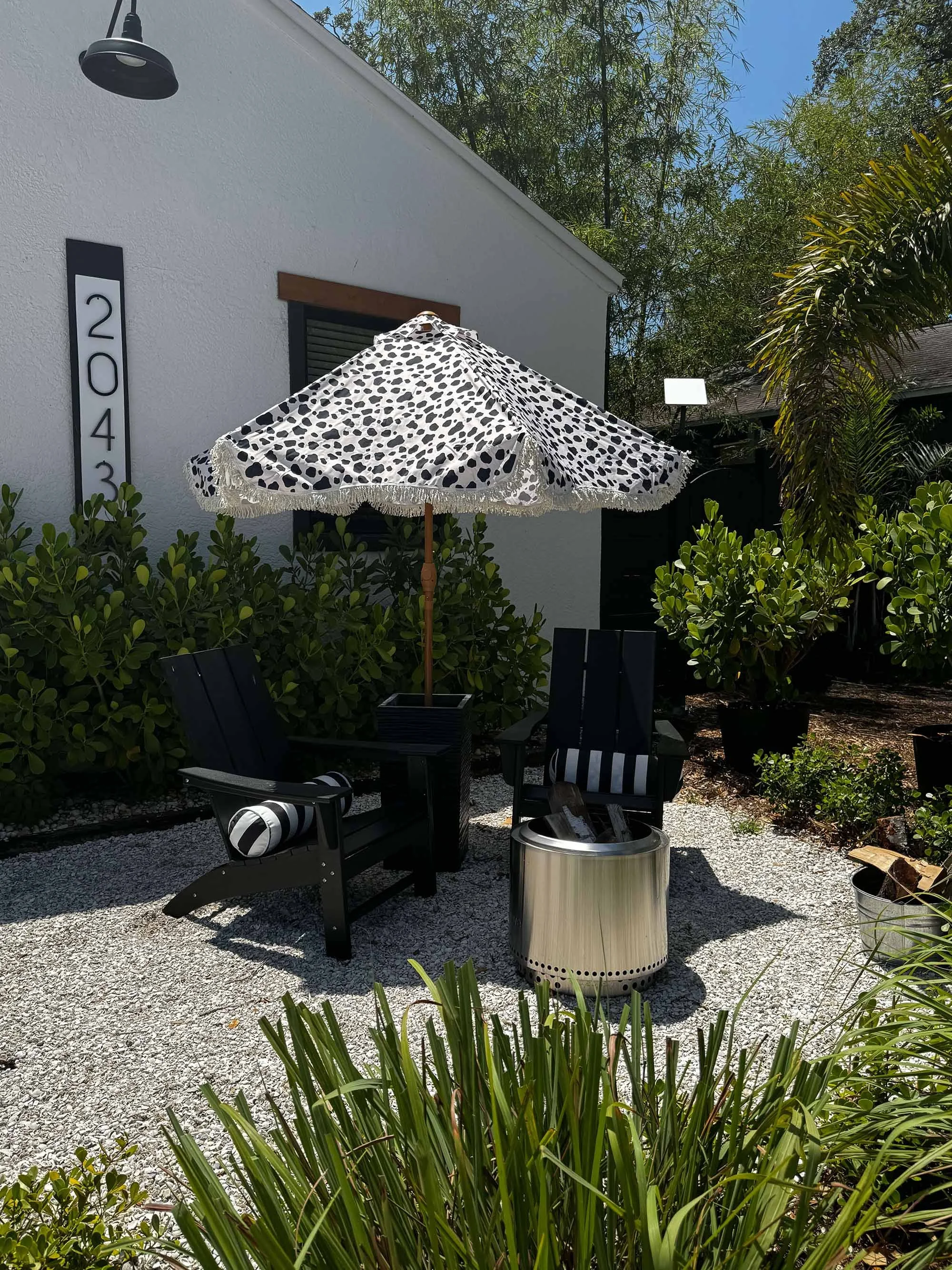 Outdoor patio area with two black chairs featuring black and white striped cushions, a black side table, a large umbrella with a black and white pattern, a silver fire pit, and lush green bushes, next to a white house.