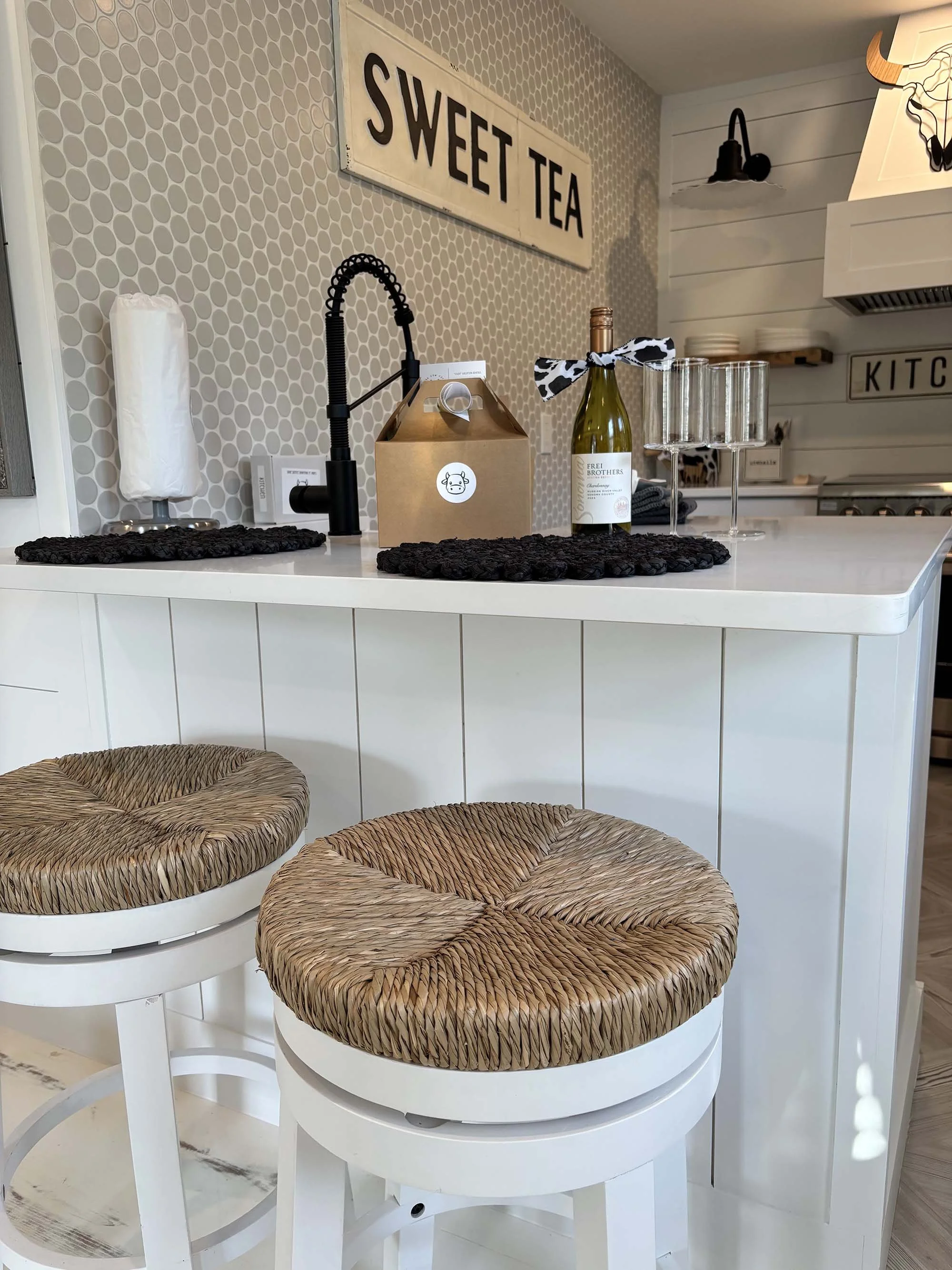 Close-up of a modern kitchen island with two woven rattan stools in the foreground. The island features a paper towel roll, a black kitchen faucet, a takeout box, a wine bottle with a bow, and empty glasses. There are wall decorations including a lar