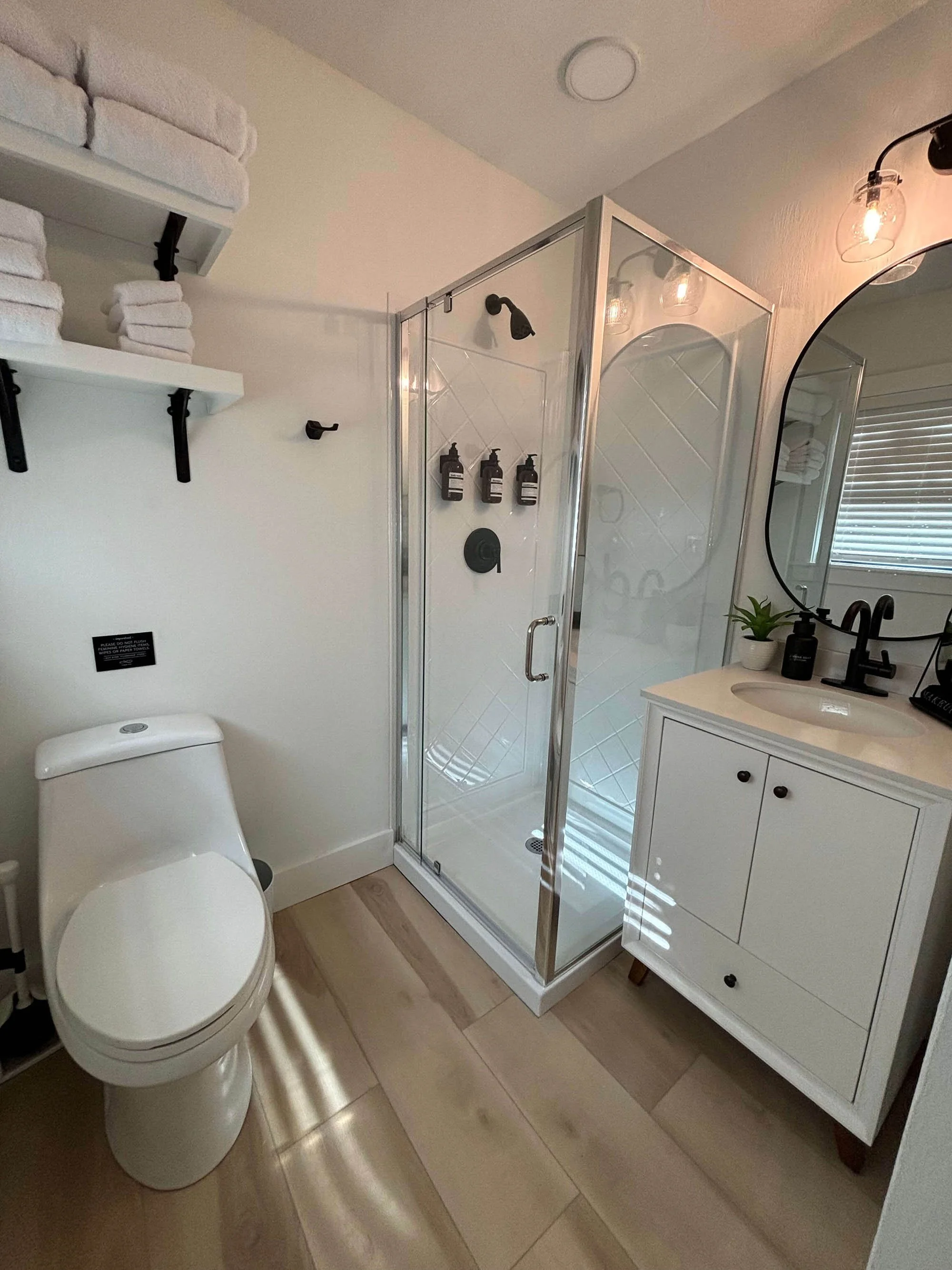 Modern bathroom with a white toilet, glass shower enclosure with black fixtures, white vanity with a black faucet, mirror, and a small plant on the sink. Shelves with white towels above the toilet, wooden floor, and a window with blinds.
