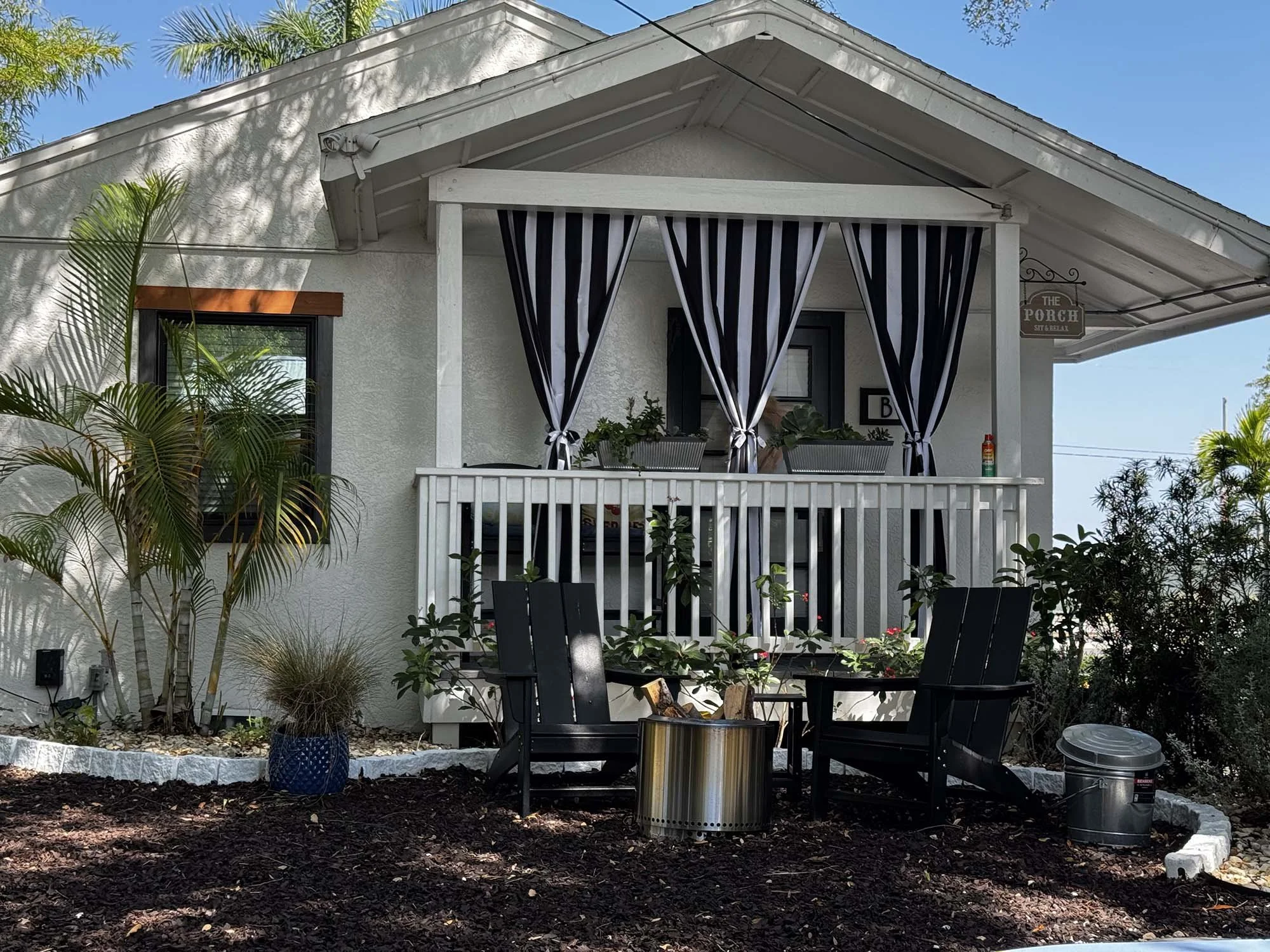 Front porch of a house with black and white striped curtains, overlooking a small garden area with two black chairs and a metal fire pit, surrounded by plants and trees.