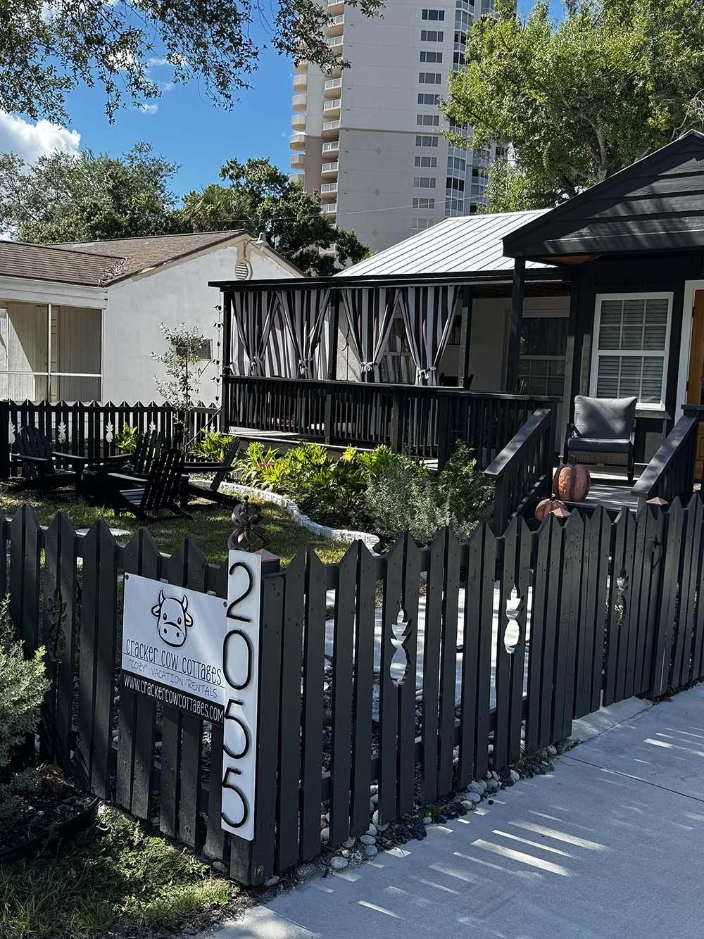 Small garden and patio area enclosed by a black wooden fence, with Adorable Cracker Cow Cottages sign. Modern buildings and tall trees in the background, with sunny weather and clear blue sky.