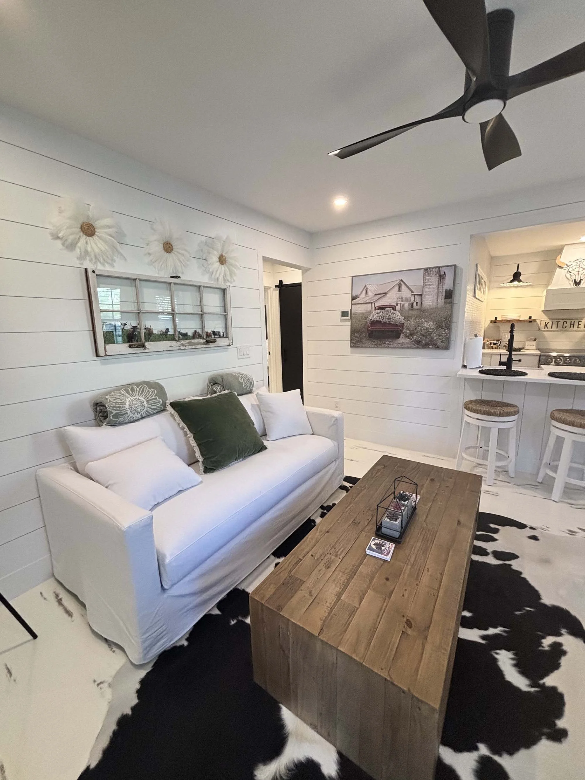 Living room with white shiplap walls, a white sofa with multiple pillows, a rustic wooden coffee table, black and white cowhide rug, wall art, and a ceiling fan, with a view into the kitchen area.