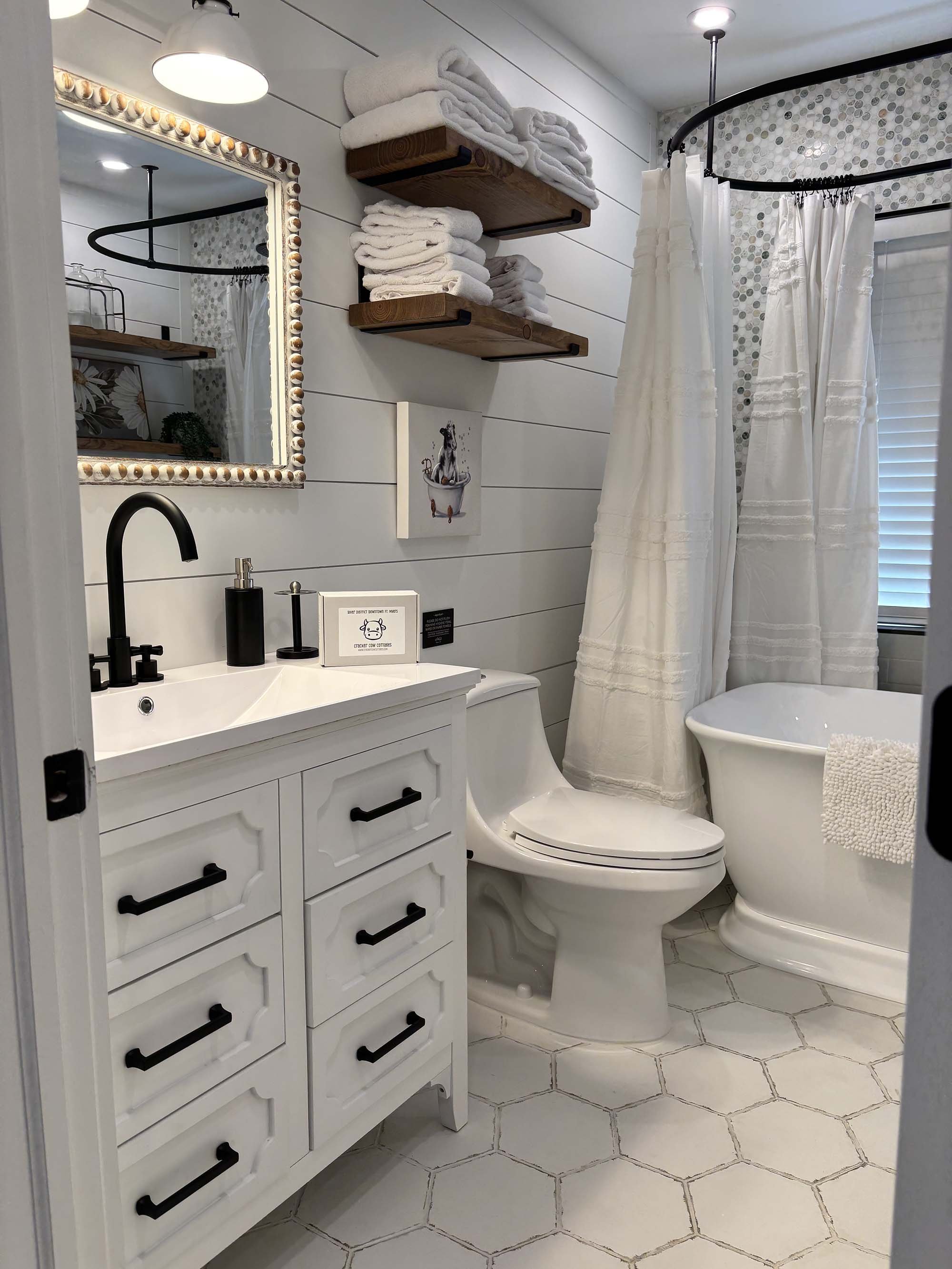 A small, modern bathroom with white shiplap walls and a hexagon tiled floor. On the left, there is a white vanity with black hardware, a black faucet, and a black soap dispenser. Above the vanity, a mirror with a decorative frame reflects the ceiling