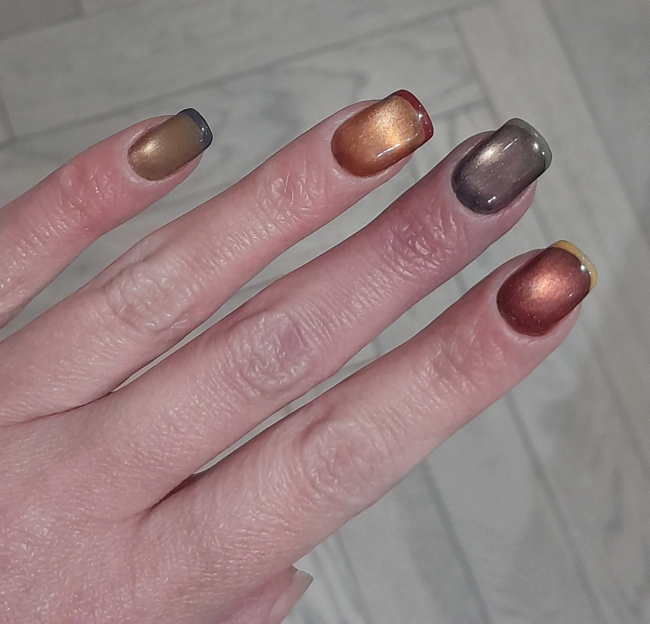 A hand with five fingers, each painted with metallic nail polish in different colors.