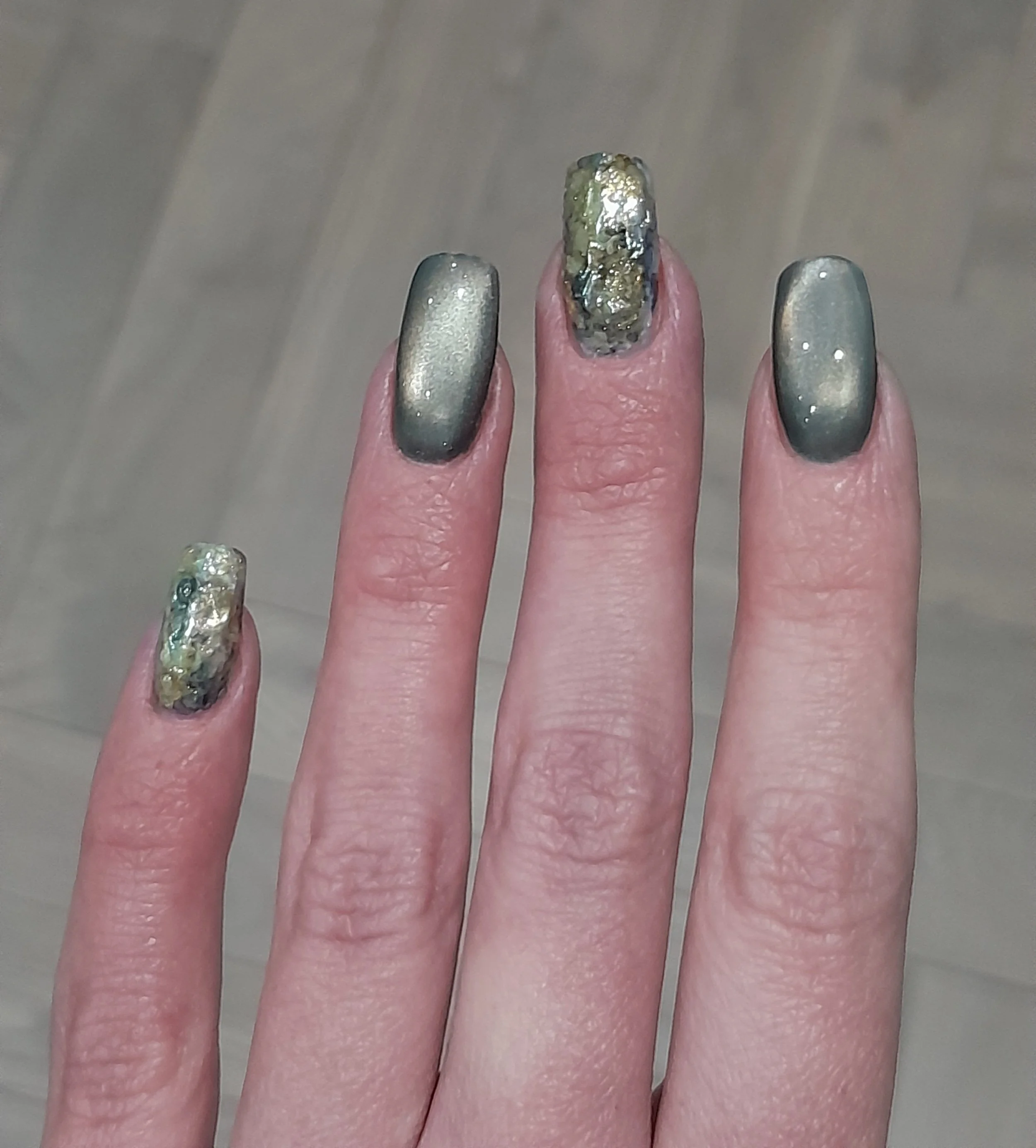 Hand with decorated fingernails, some with a metallic silver gradient and others with a textured, multi-colored design with gold and silver accents.