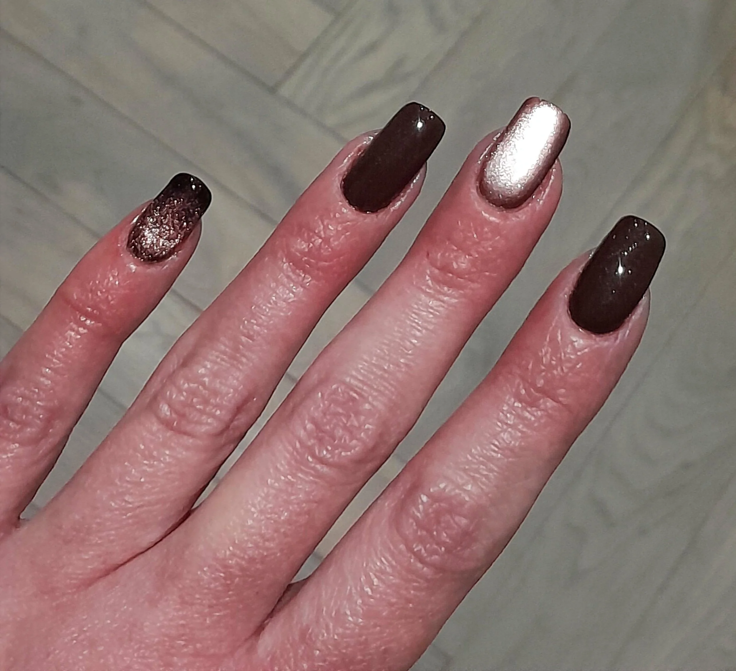 Close-up of a woman's hand showing five fingers with dark-colored and metallic nail polish on her nails.