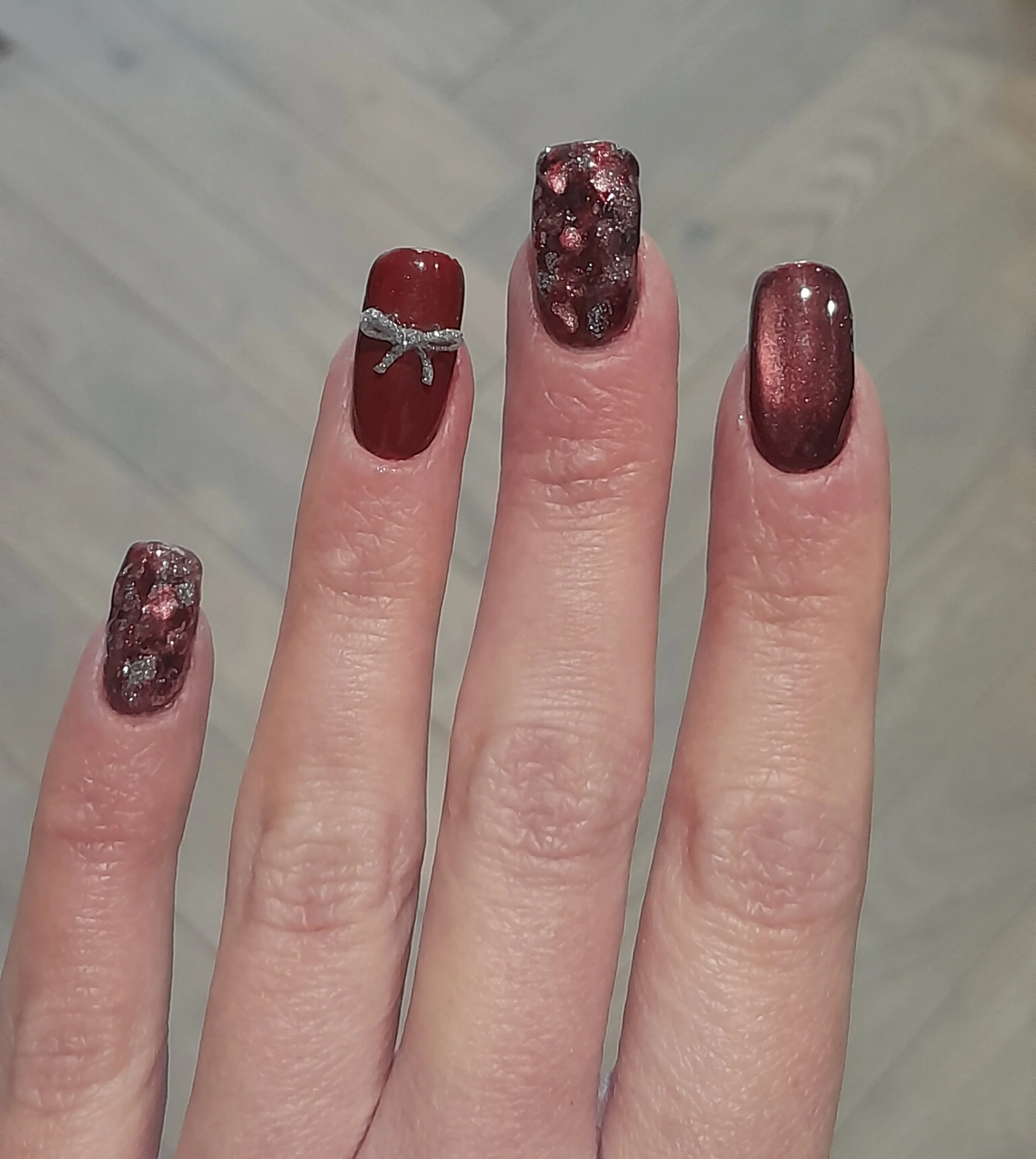 Manicured nails painted in a deep red color with glitter accents, one with a silver embellishment, on a person's hand with visible skin texture and natural light.