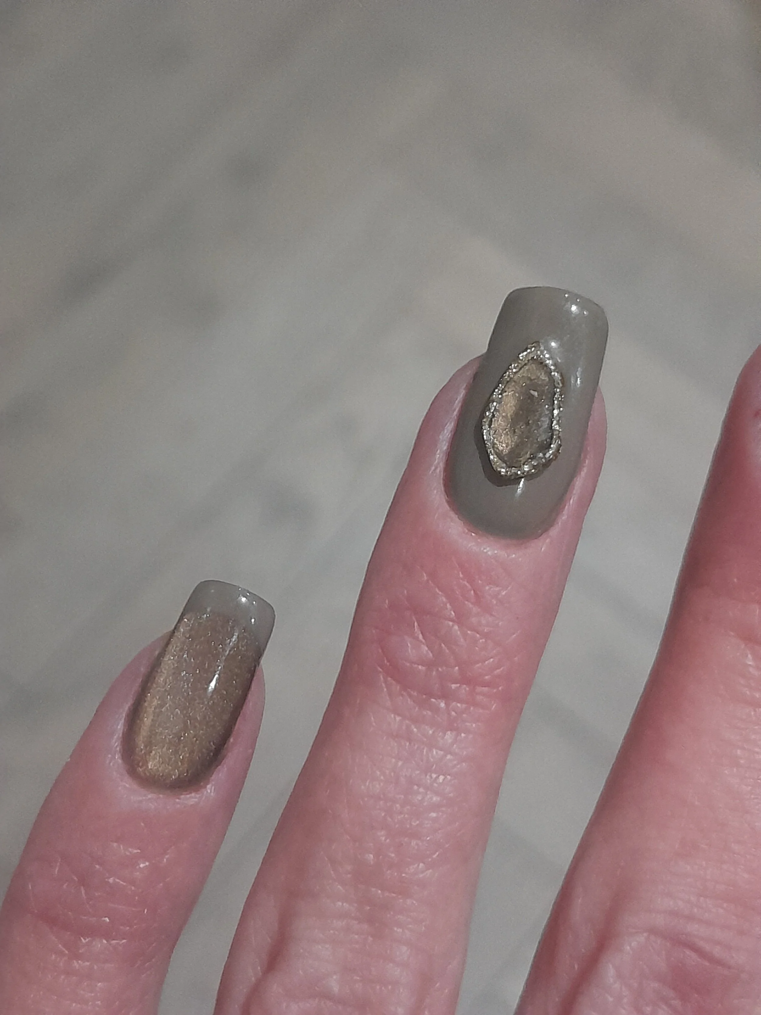 Close-up of a hand with nails painted in glossy taupe color, featuring an oval metallic embellishment with a gold and silver design on one nail.