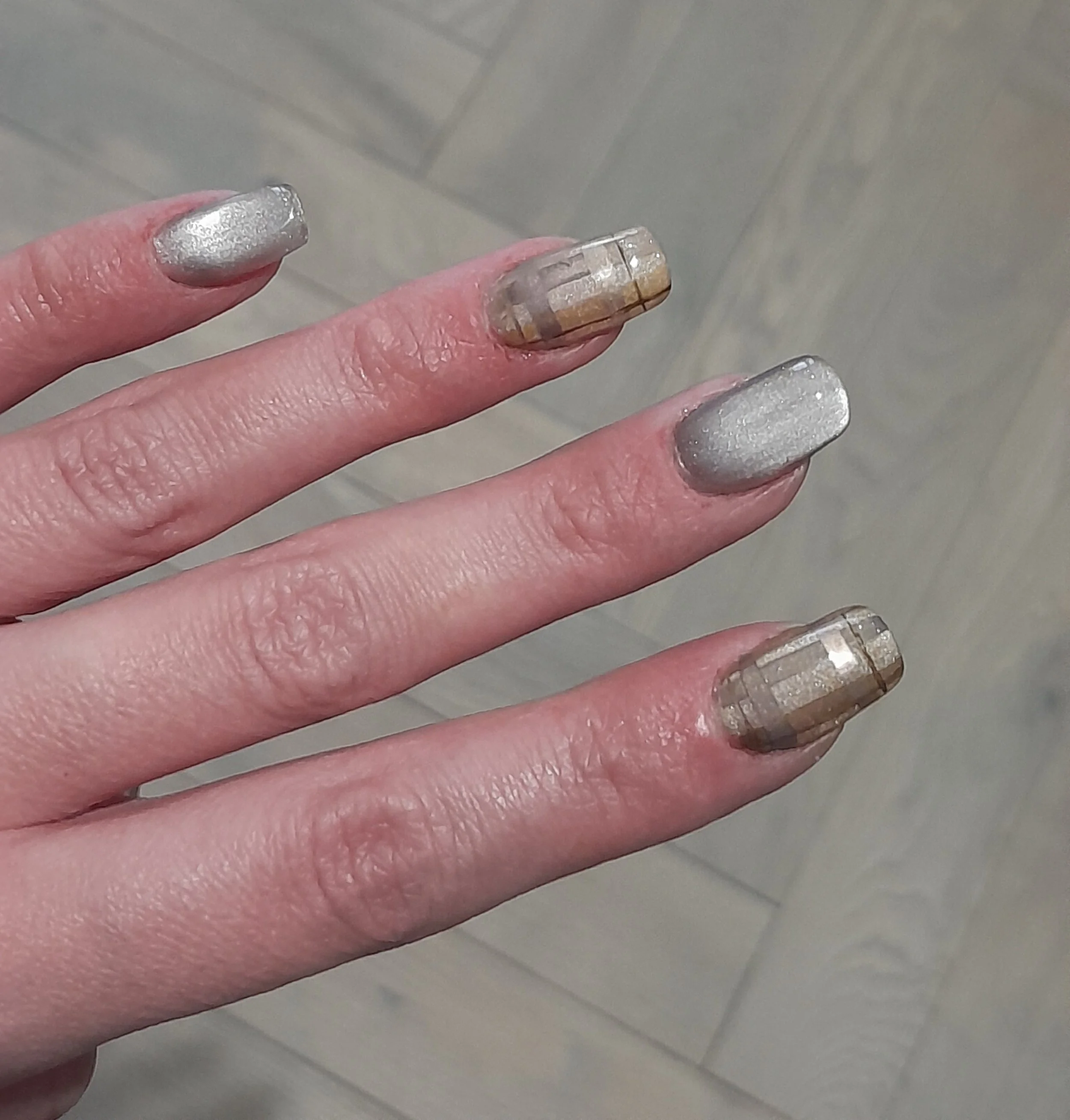 Close-up of fingers with metallic silver and beige plaid nail polish against a wooden floor background.
