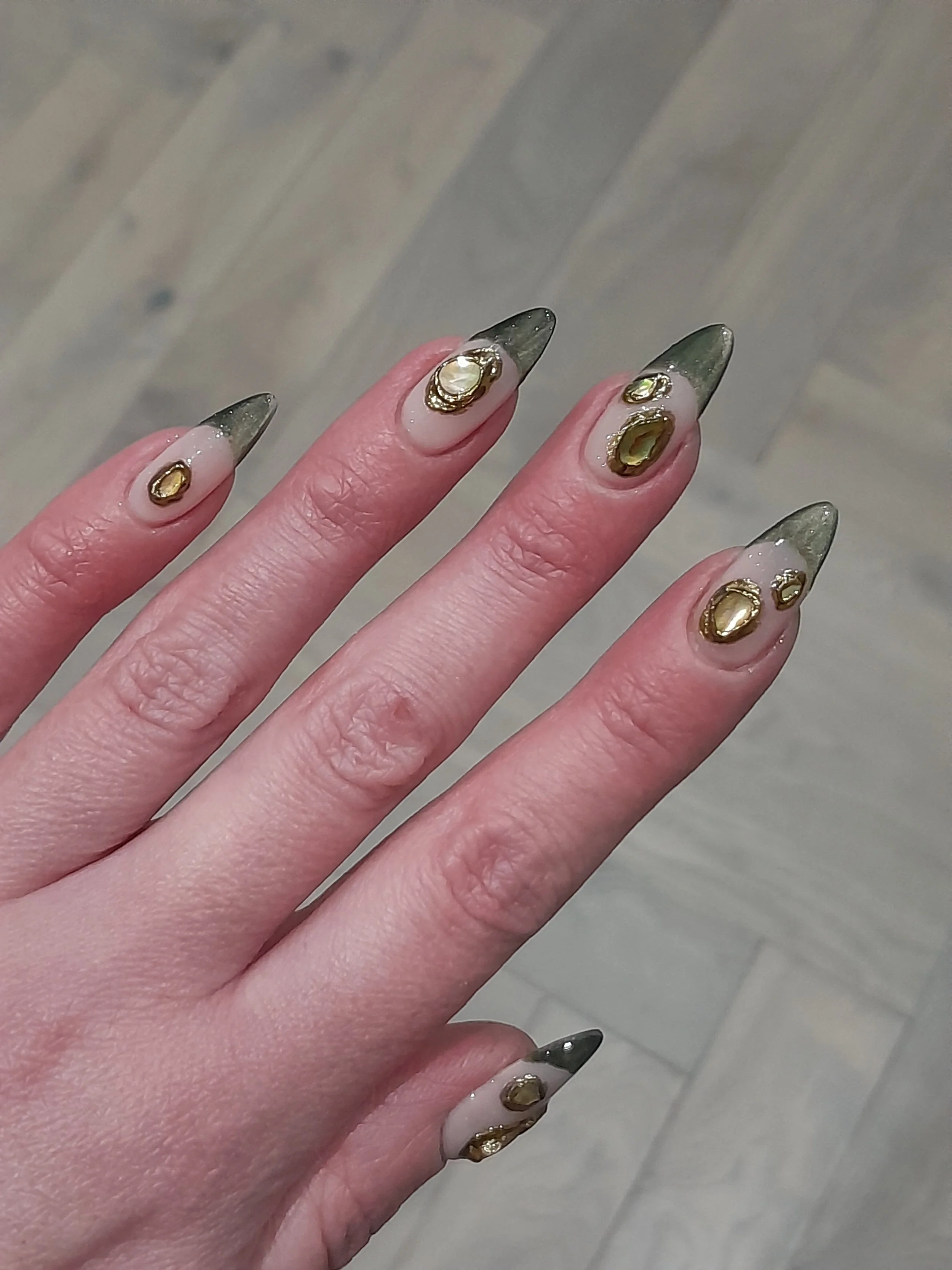Close-up of a person's hand with long, almond-shaped nails featuring a black and clear gradient design with gold embellishments.