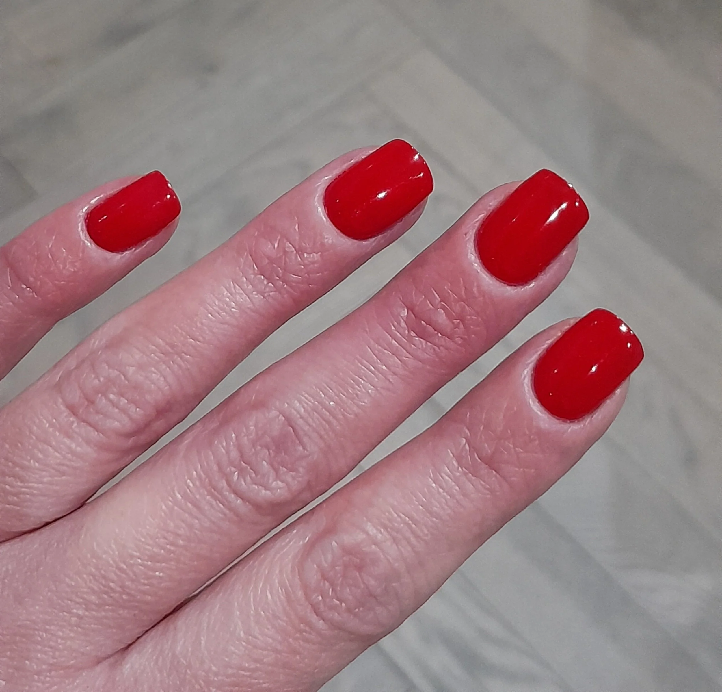 Close-up of a hand with four fingers visible, each with red-painted nails. The background shows a light-colored wooden floor.