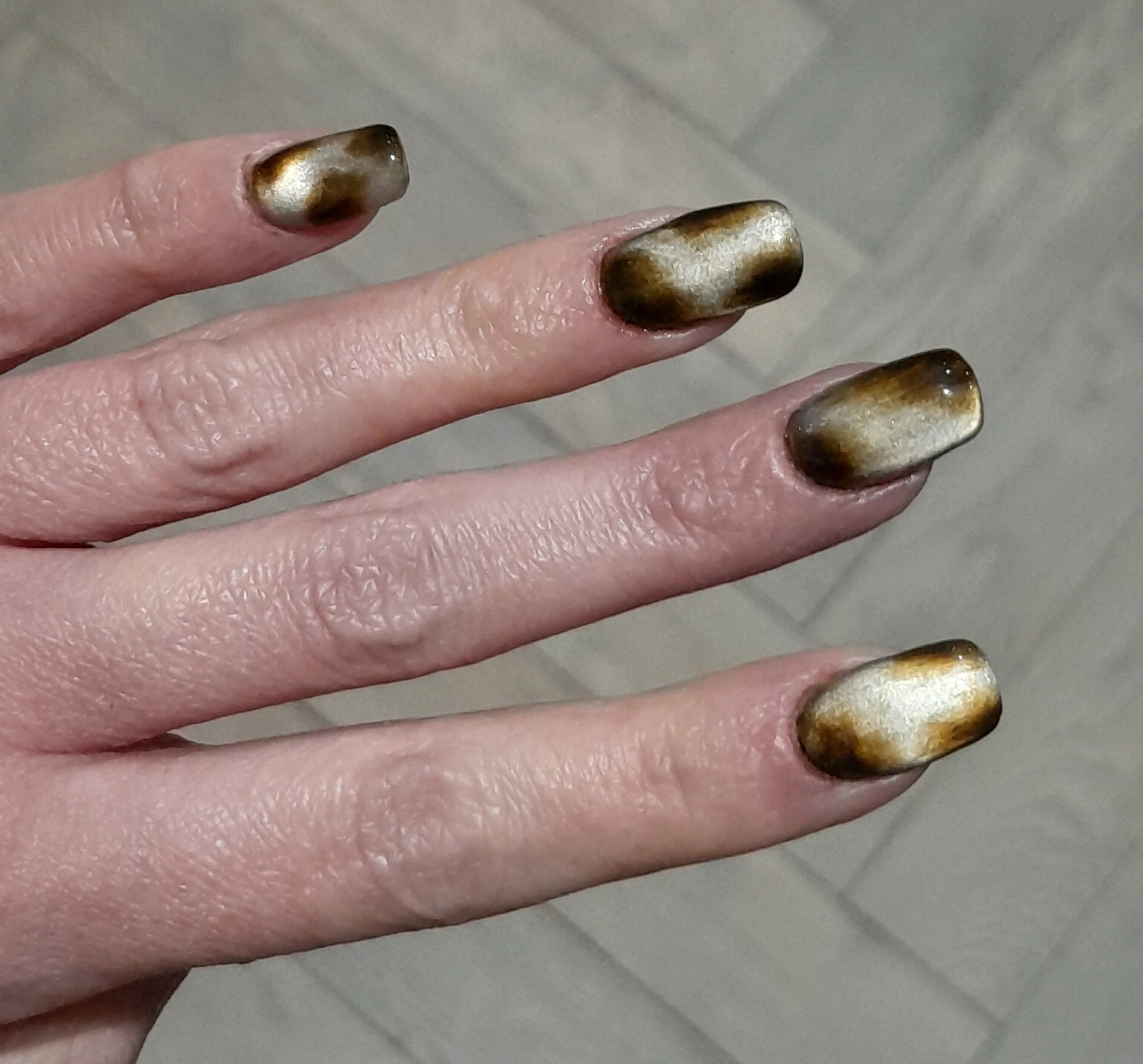 Hand showing fingers with metallic nail polish that has a black, gold, and silver gradient design.