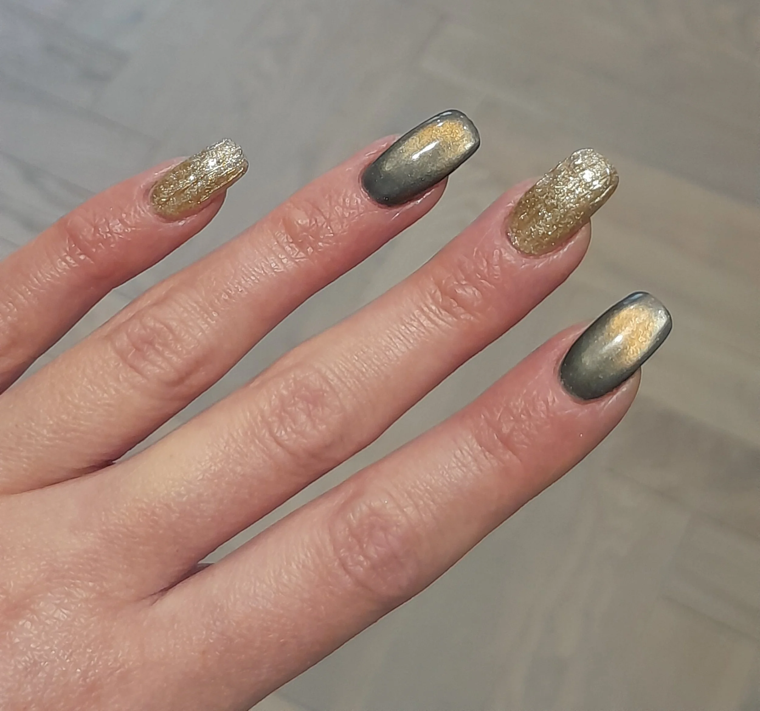 Close-up of a hand with nails painted in metallic gold and black gradient with a textured gold accent on some nails.