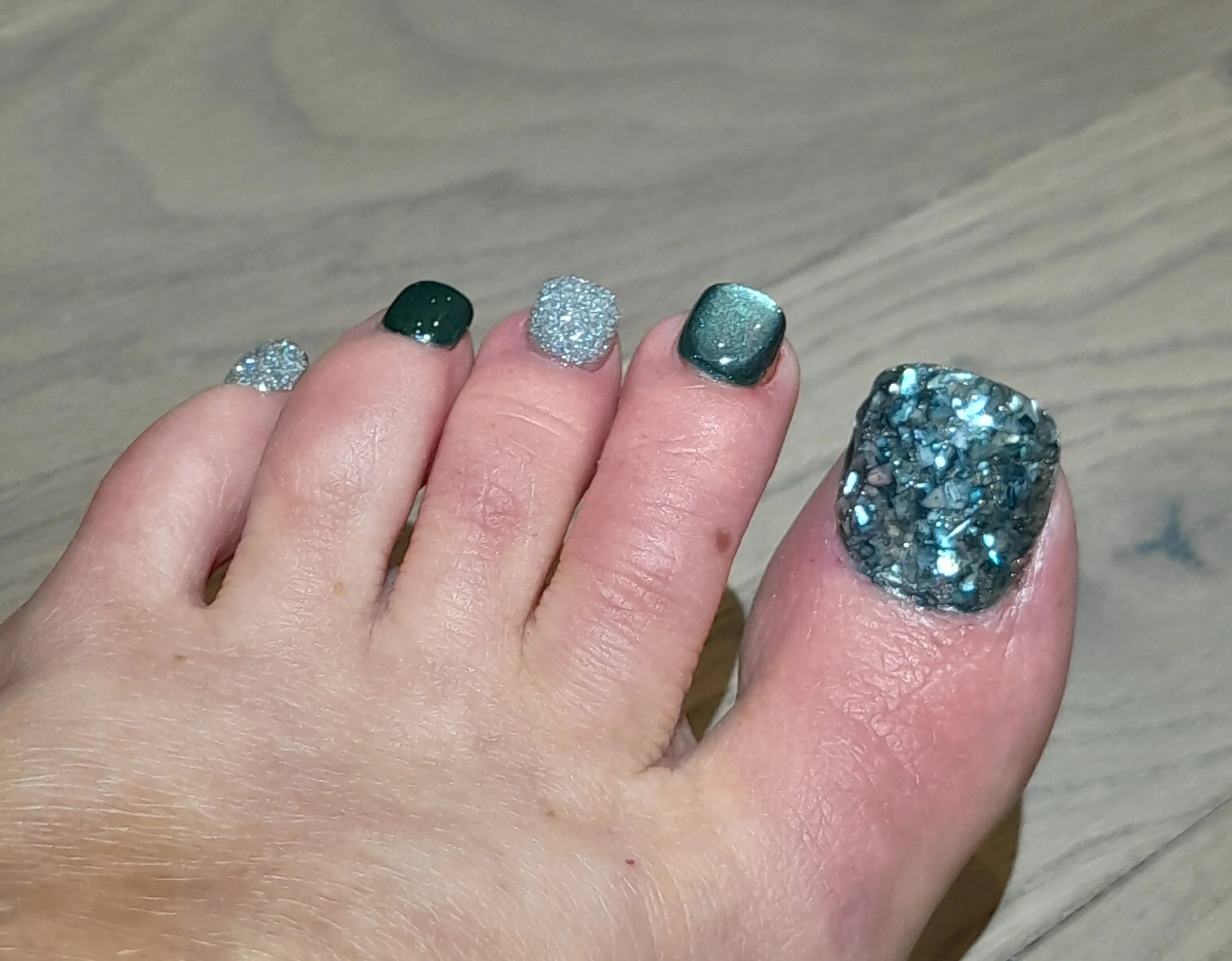 Close-up of a person's hand with manicured nails featuring sparkly and metallic nail polish in shades of green, silver, and black-gray, with the thumb nail covered in a dark, glittery, textured polish.