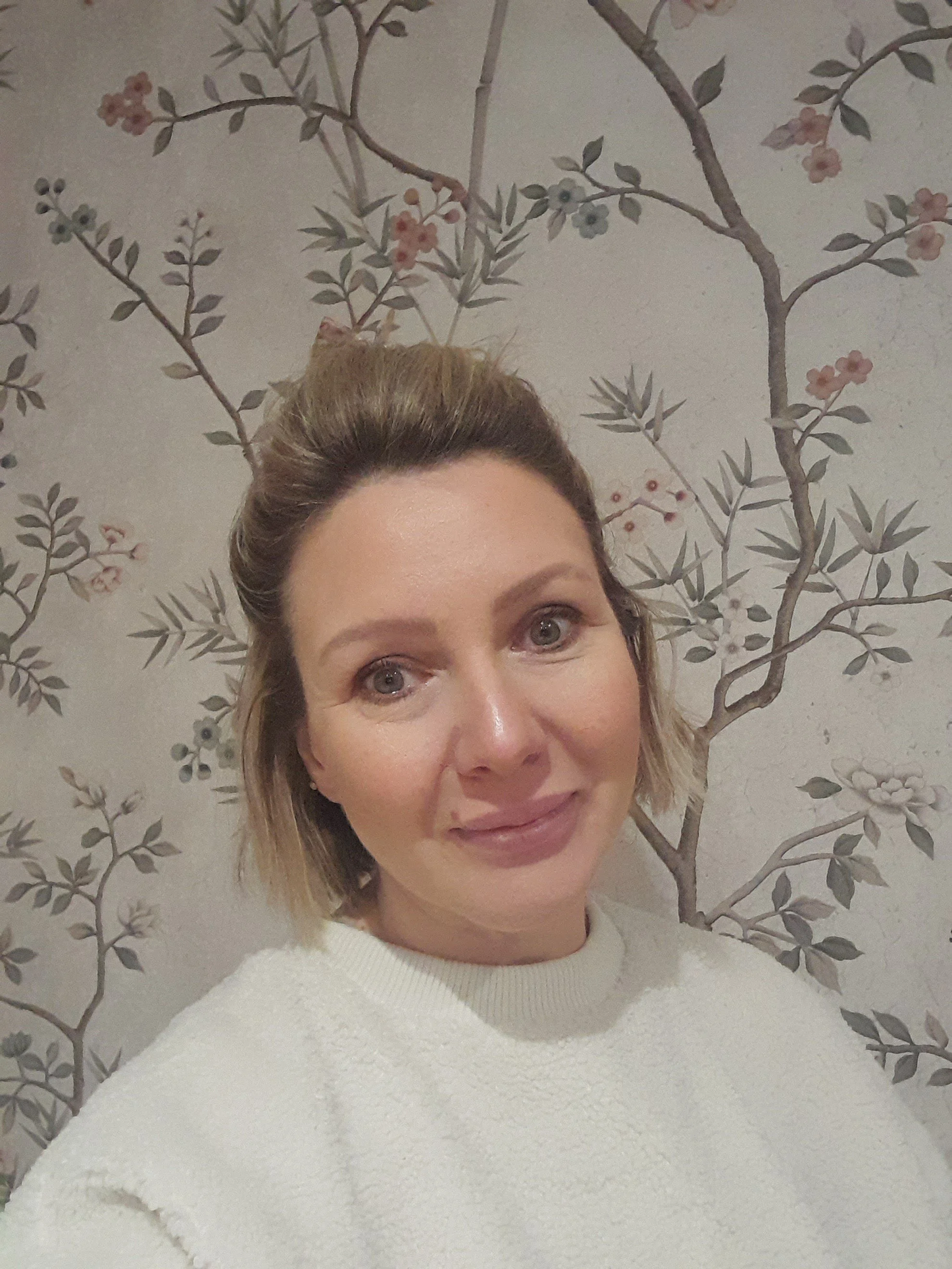 A woman taking a selfie in front of a wall with a floral tree pattern wallpaper.
