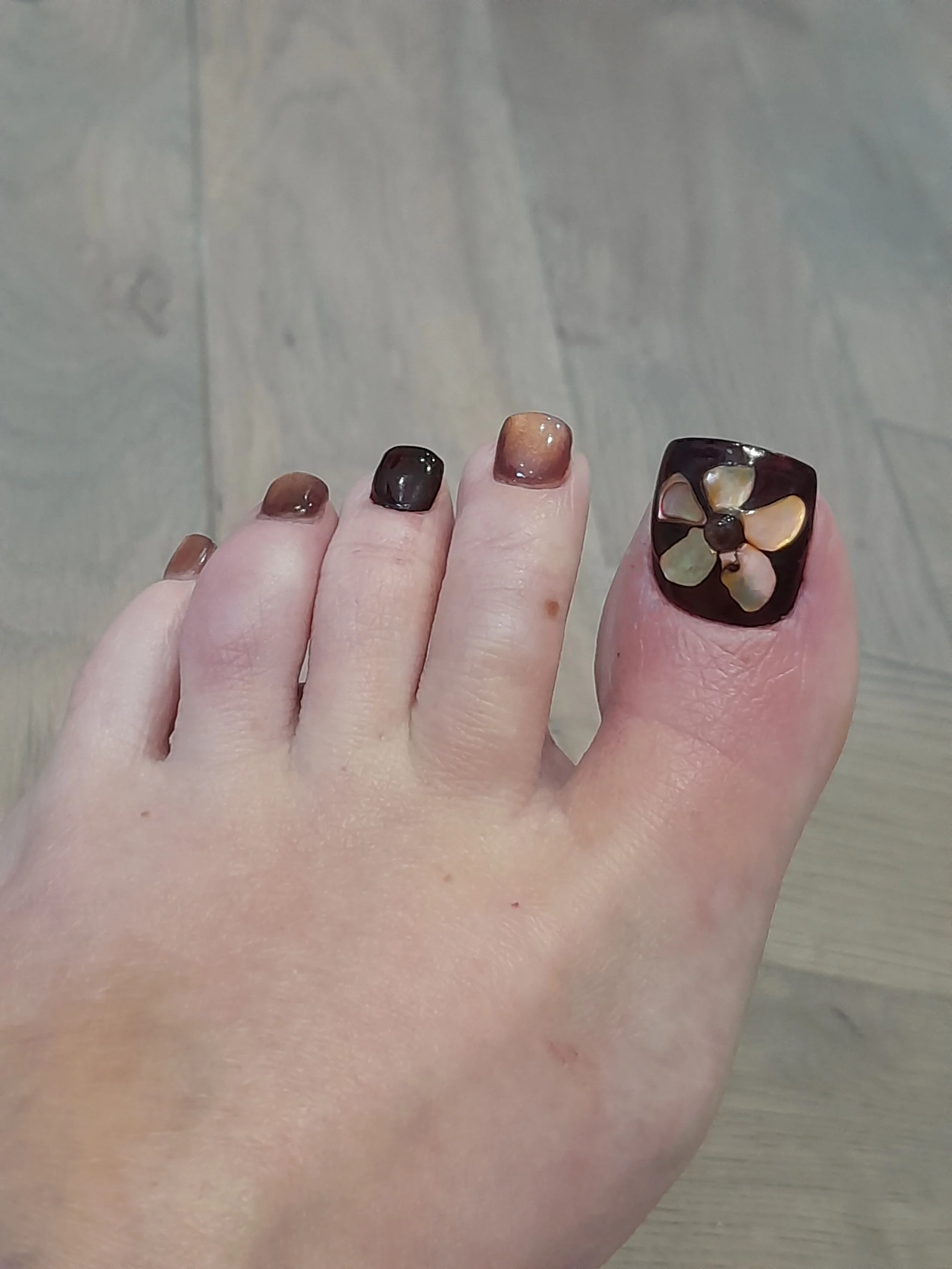 A person's foot with toenails painted in a mix of dark brown, gold, and beige, with the big toe featuring an inlaid flower design made of shell or mother-of-pearl in a black square nail polish.