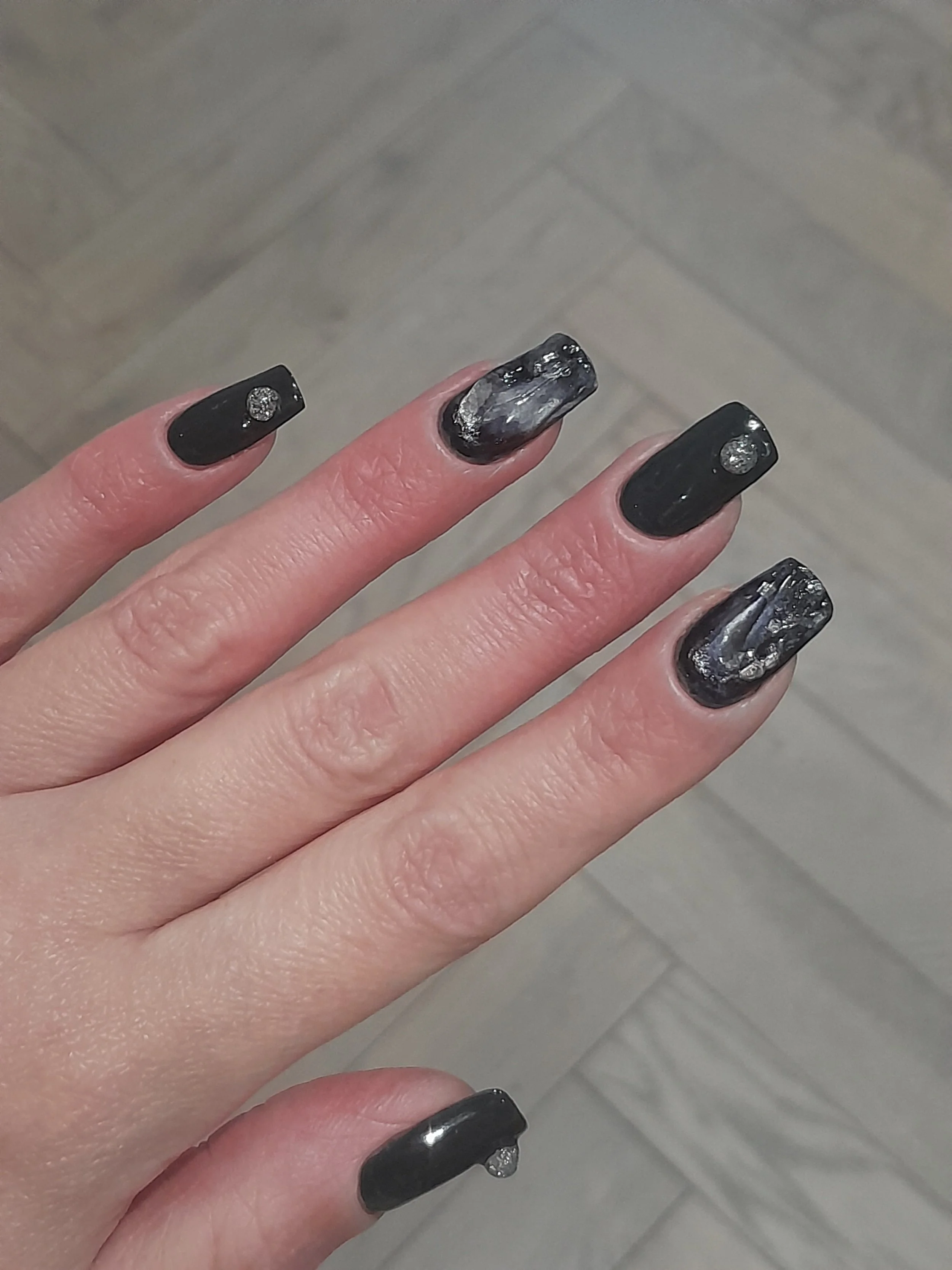 Close-up of a hand with black and silver marble patterned nail polish and small rhinestones on the index, middle, and pinky fingernails.