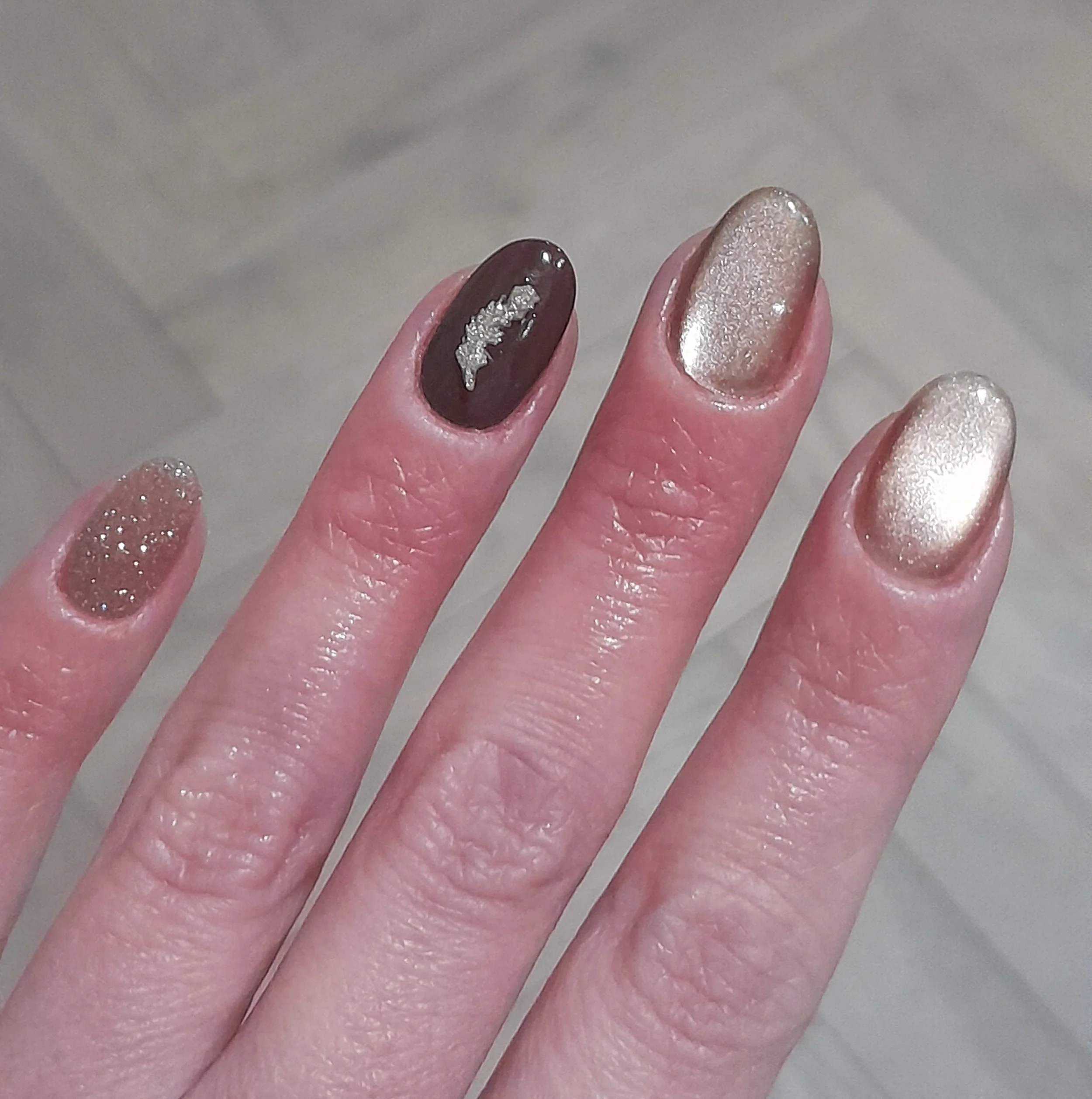 Close-up of a hand with five fingers, each with a different metallic or glittery nail polish, including shades of gold, bronze, and dark brown with silver embellishments.