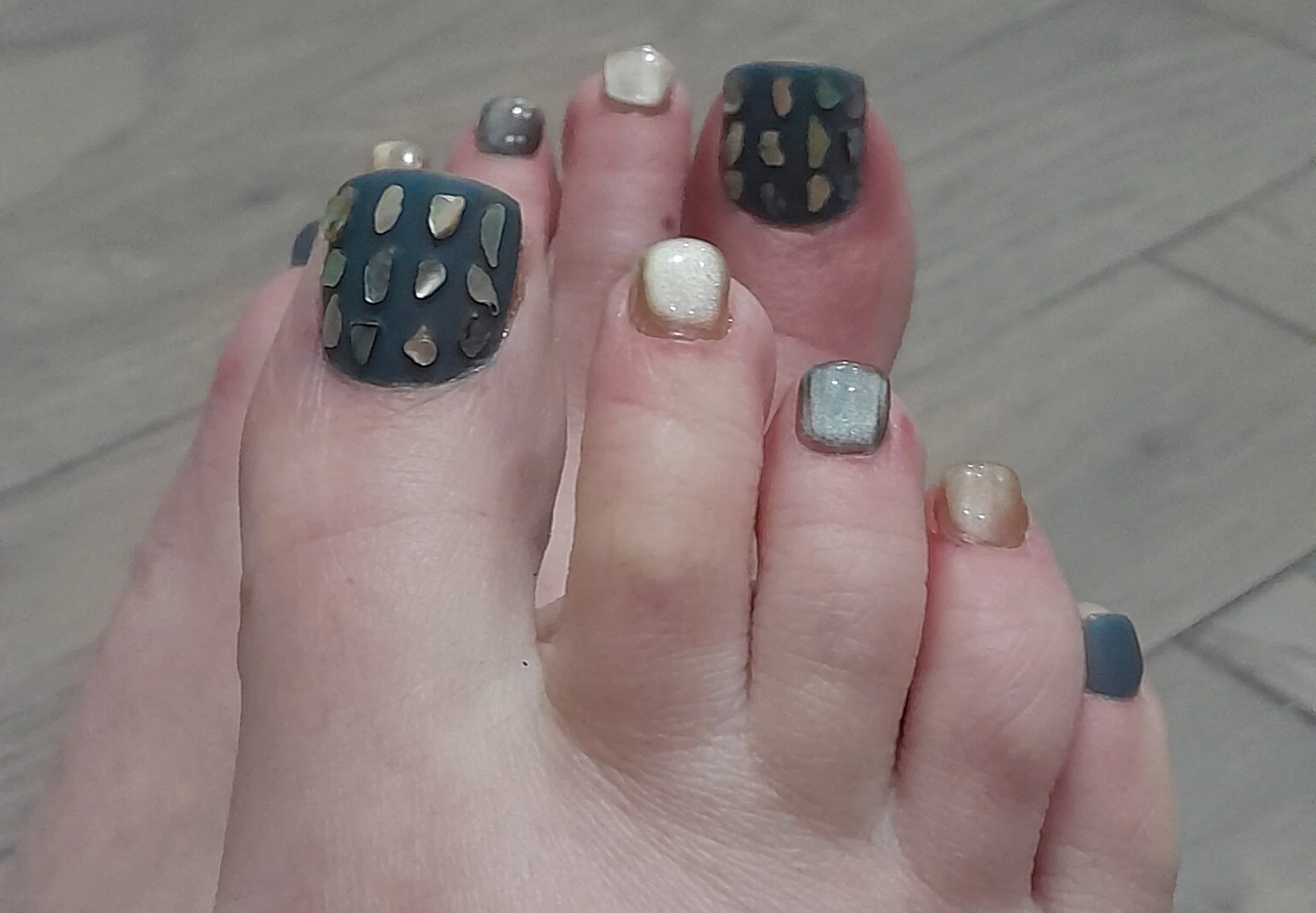 Close-up of a person's foot showing toenail polish with various colors and designs, including glitter, metallic, and painted patterns.